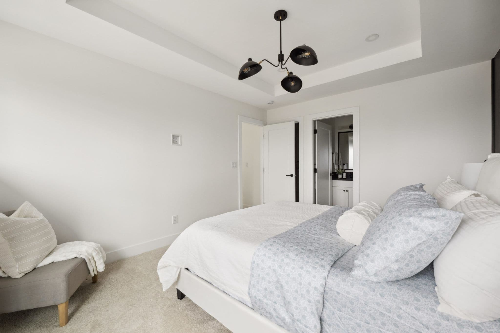 Water Tower Hill – A bright, neutral bedroom featuring a neatly made bed dressed in white and light blue patterned linens, complemented by light carpeting and white walls with a tray ceiling. A modern black three-bulb chandelier hangs overhead, while an open doorway in the background reveals an ensuite bathroom.