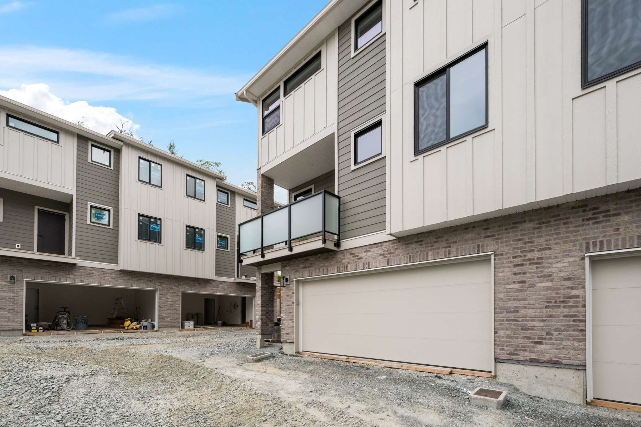 Water Tower Hill – The exterior rear of newly constructed three-story townhomes, featuring brick ground-level garage facades, modern vertical board-and-batten siding above, and frosted glass balcony railings. The foreground shows an unpaved gravel laneway under a bright, partly cloudy blue sky, indicating near-completion construction.