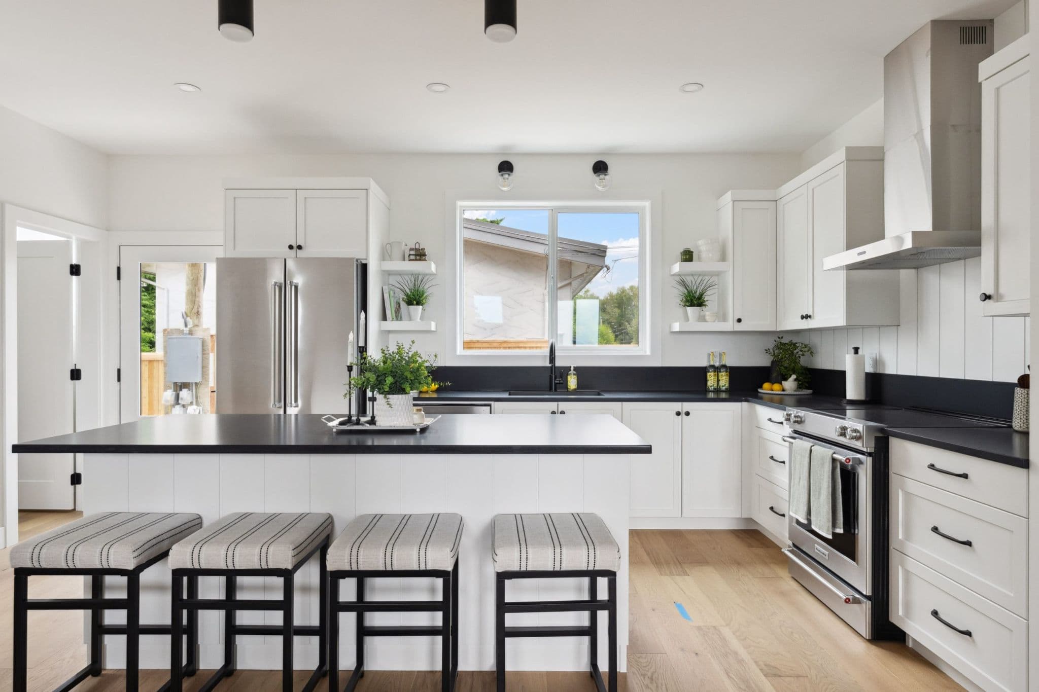 Water Tower Hill – A bright, modern farmhouse-style kitchen features crisp white shaker cabinetry, contrasting black countertops, and light hardwood floors. A large central island with four striped upholstered barstools sits in the foreground, flanked by stainless steel appliances and a window looking out onto a residential yard.