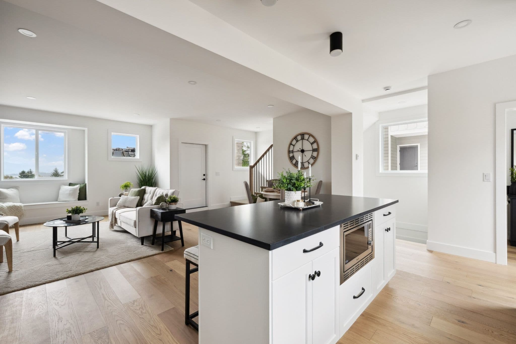 Water Tower Hill – A bright, open-concept main floor features a white kitchen island with a black countertop and built-in microwave in the foreground, flowing into a furnished living room. Light wood flooring runs throughout the space, which is illuminated by daylight from multiple windows showing clear blue skies outside.