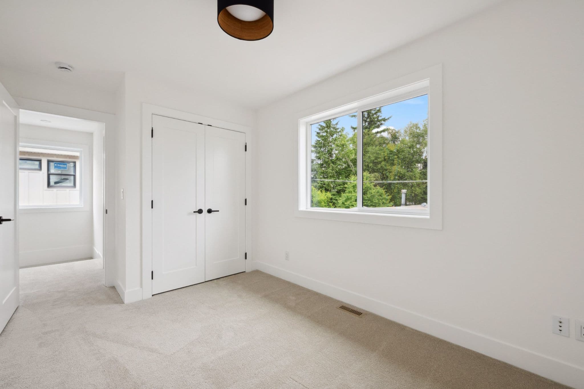 Water Tower Hill – An empty bedroom featuring beige carpeting, crisp white walls, and a double-door closet with black hardware. A large window brings in bright daylight and looks out onto lush green treetops under a clear blue sky.