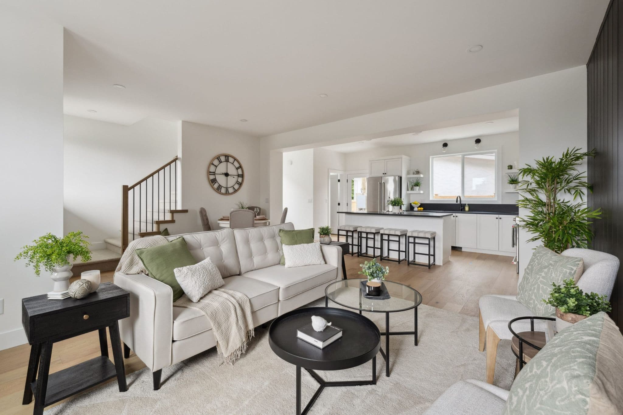 Water Tower Hill – A bright, open-concept living area seamlessly connects to an adjacent dining nook and modern kitchen with a breakfast bar. The space is styled with light wood flooring, a cream tufted sofa with sage green accents, nested coffee tables, and contemporary white cabinetry.