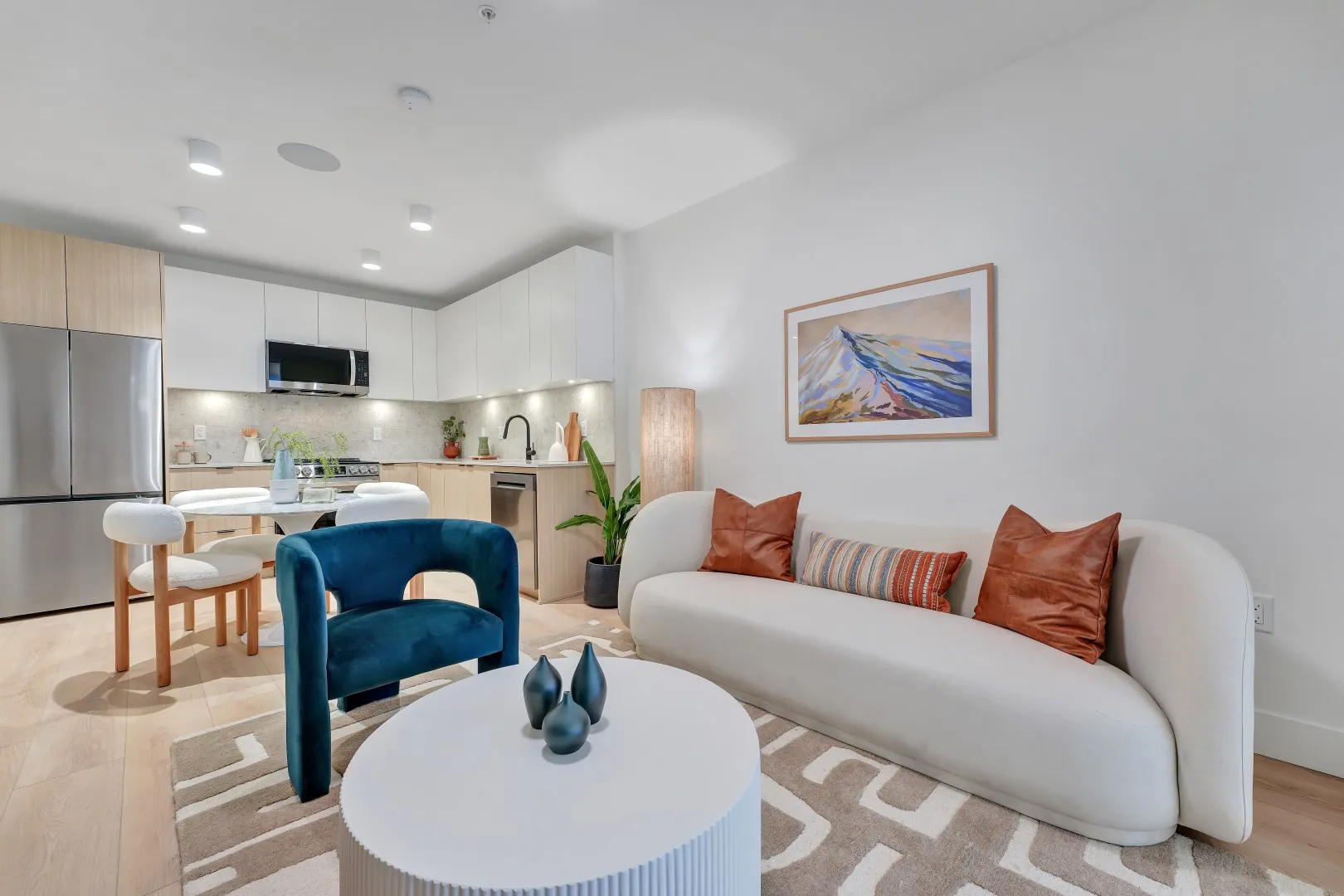 Jethro – Living room and kitchen interior with a teal velvet accent chair, cream sofa with rust pillows, and a mountain-themed framed print.