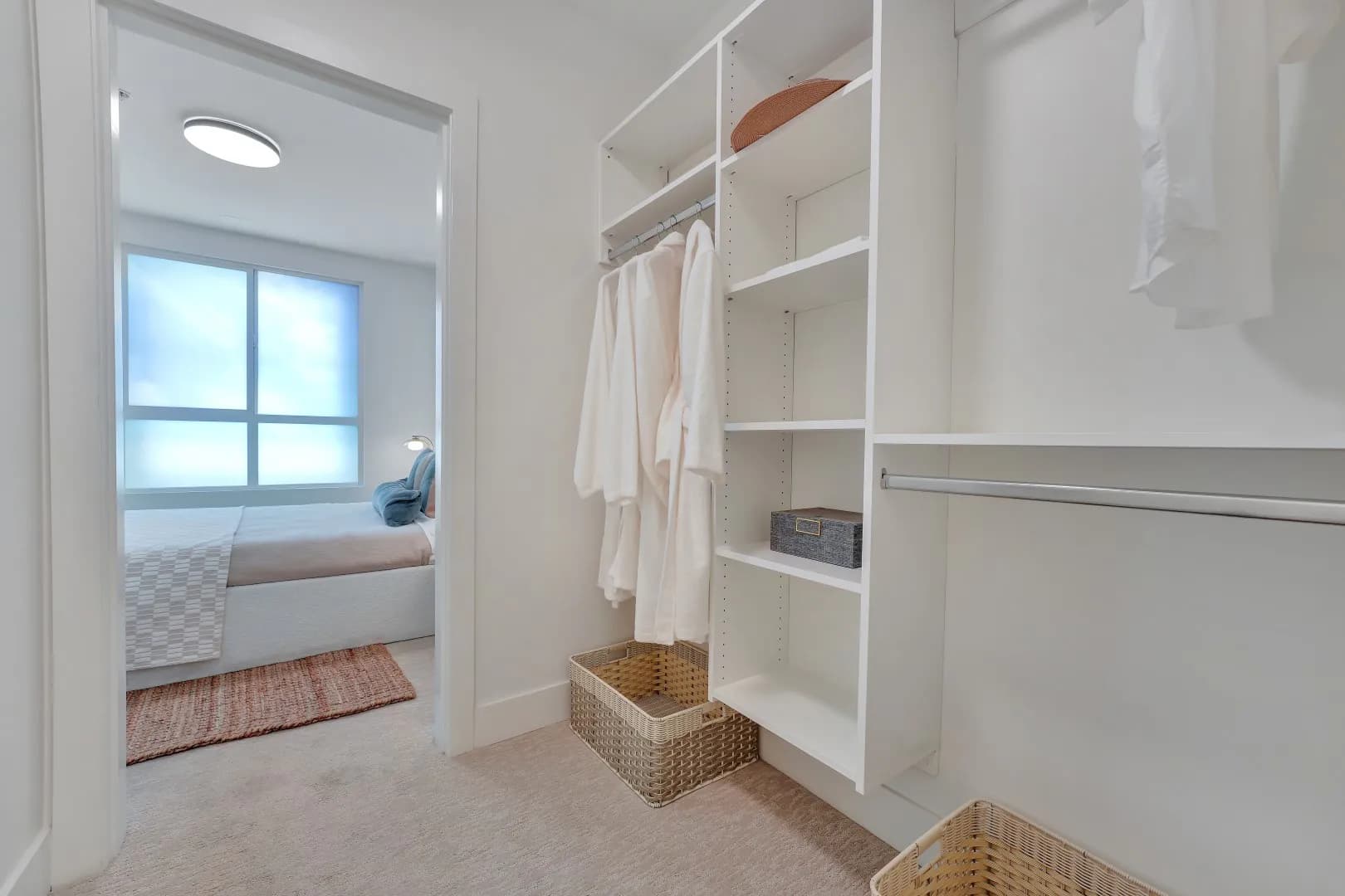 Jethro – Walk-in closet with white shelving, hanging robes and a shirt, woven storage baskets, seen through to the bedroom beyond.