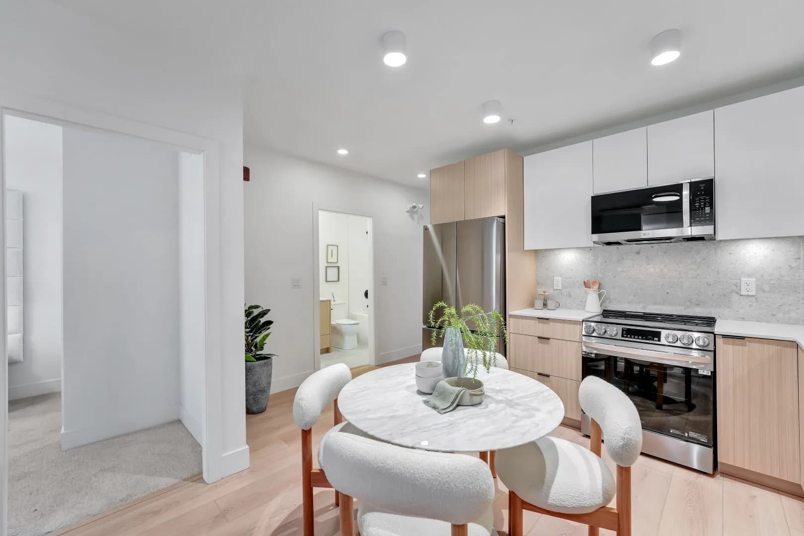 Jethro – Kitchen and round dining table area with stainless refrigerator and range, white and wood cabinetry, and a glimpse into an adjoining bathroom.