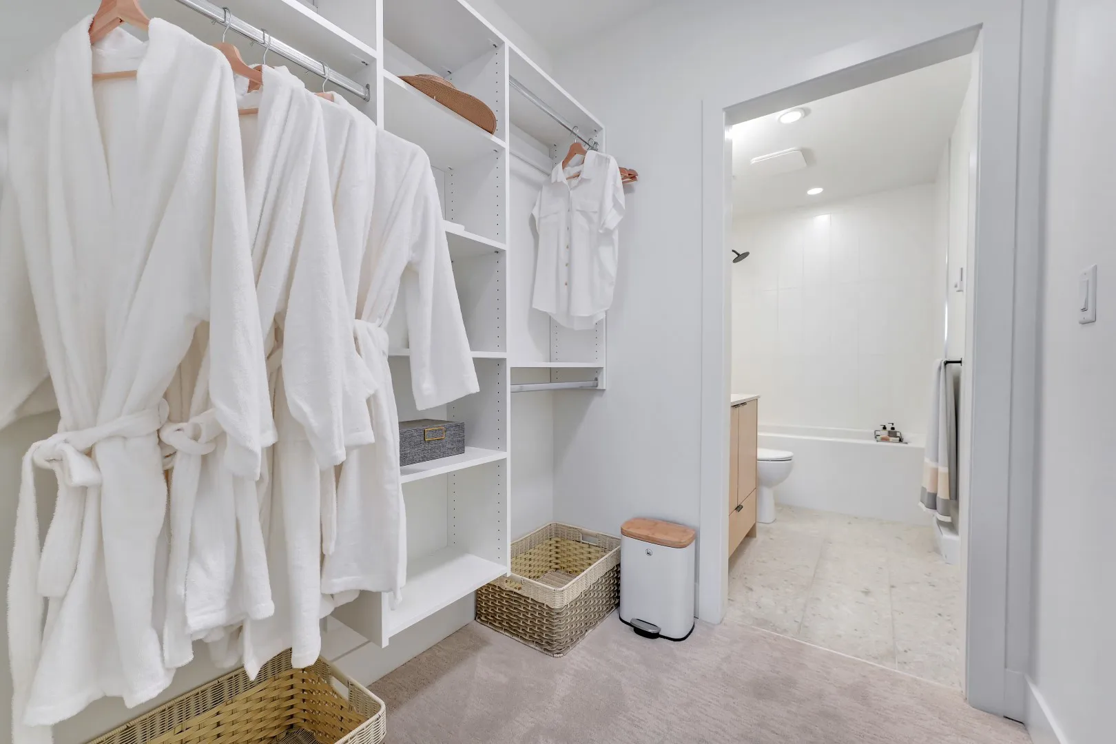 Jethro – Closet interior with hanging white robes and shirts, woven baskets, and an open doorway into a bathroom with a soaker tub.
