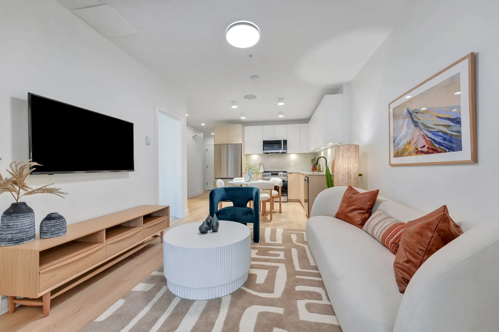 Jethro – Open-concept living room with a wall-mounted TV, wood media console, cream sofa with rust throw pillows, and a view into the kitchen and dining area.