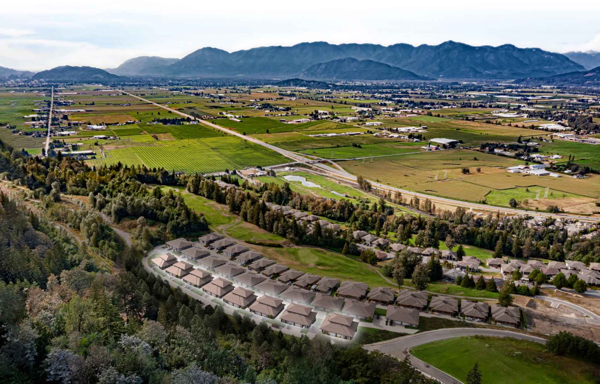 Vista Green at The Falls – Aerial drone photo of the townhome community nestled against a forested hillside with farmland, mountains, and a highway in the background.
