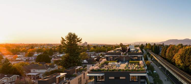 Clive at Collingwood Condos – Aerial sunset view of a rooftop terrace with lounge furniture and greenery atop a low-rise building, with a train line and mountains beyond.