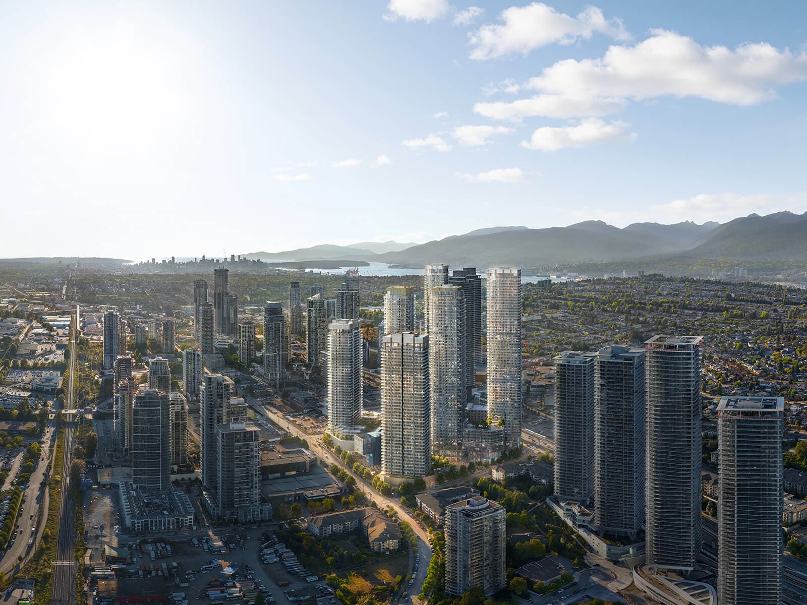 Brentwood Block – A high-angle drone shot captures a sprawling urban cityscape bathed in bright daylight, featuring a cluster of modern glass high-rise residential towers surrounded by lower-density neighborhoods. In the background, a body of water separates the city skyline from a mountain range under a clear sky with soft clouds.