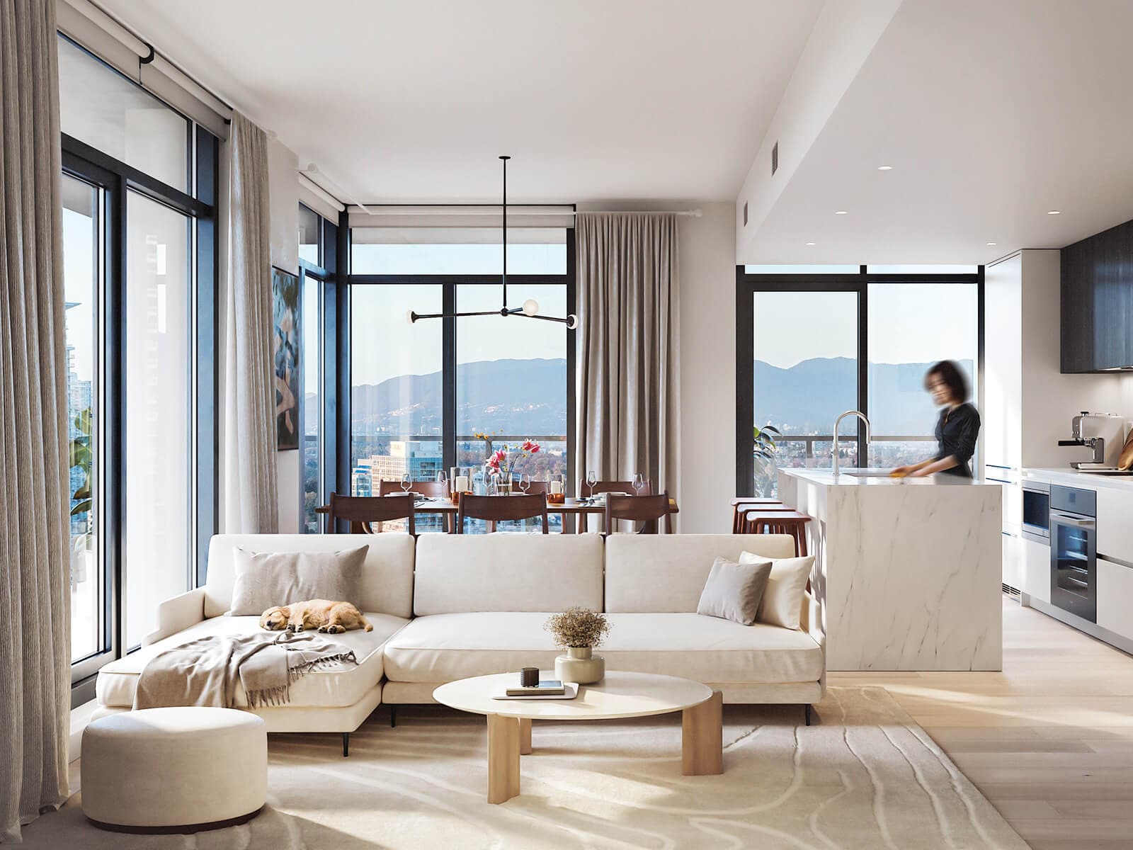 Brentwood Block – A bright, modern open-concept living area featuring a light cream sectional sofa with a dog resting on it, leading into a dining space and a kitchen with a marble waterfall island. Floor-to-ceiling windows flood the suite with natural daylight and offer expansive views of distant mountains and the city skyline.