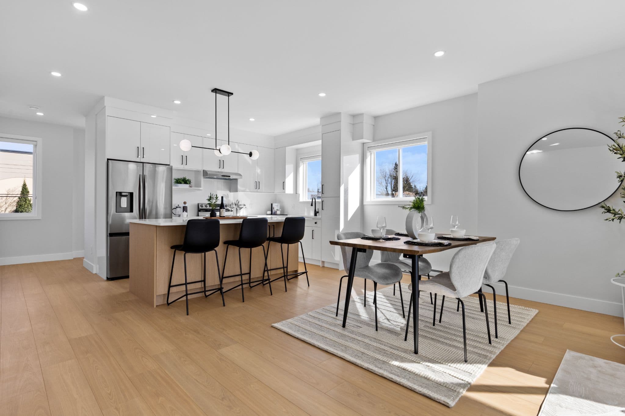 Clearbrook Commons – Modern open-concept kitchen and dining area with white cabinetry, light wood flooring, kitchen island with black bar stools, and a dining table set for four.