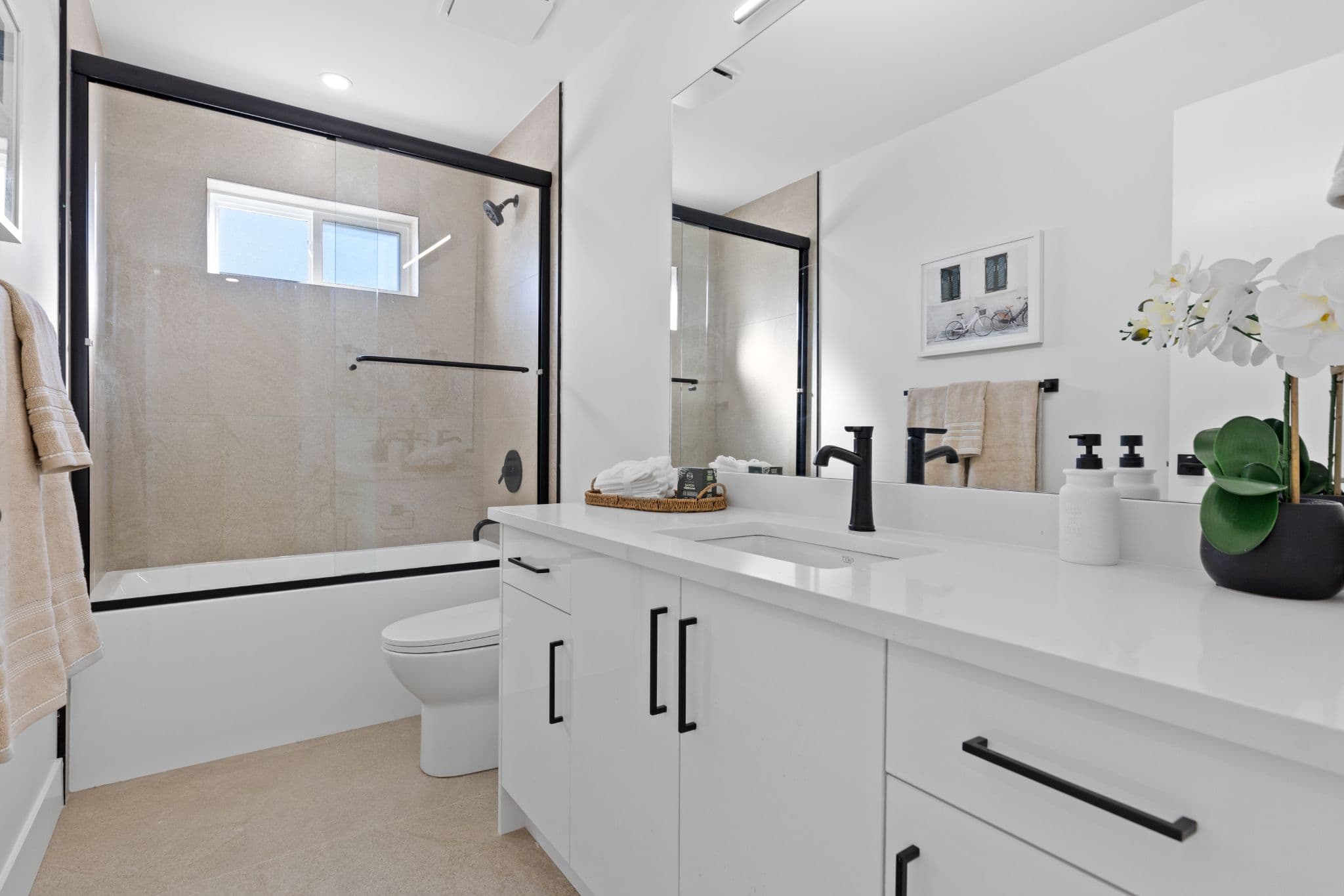 Clearbrook Commons – Full bathroom featuring a white double-sink vanity with black faucets, toilet, and glass-enclosed tub and shower combination.