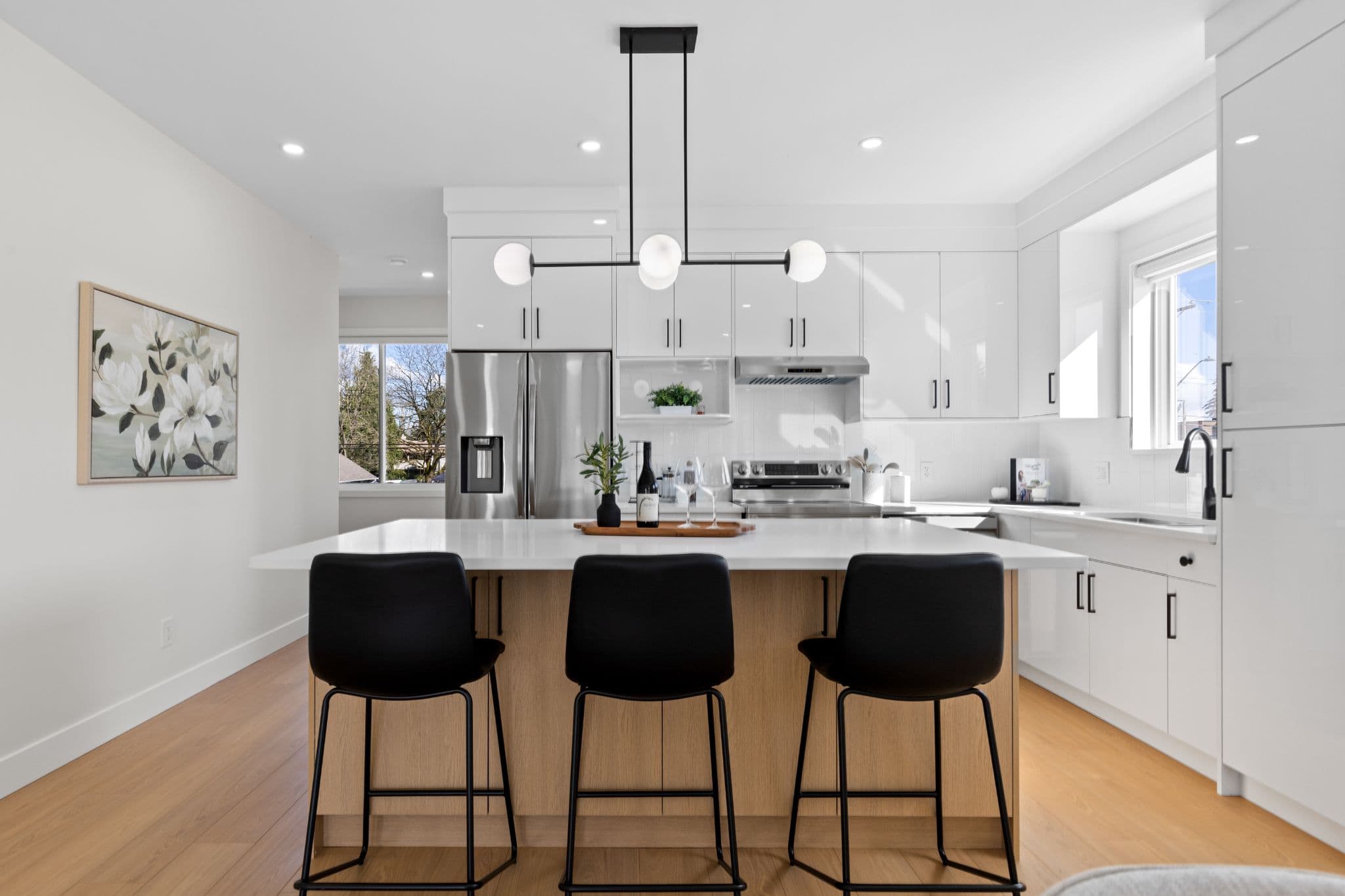 Clearbrook Commons – Kitchen island with wood trim and three black bar stools under modern pendant lights, facing white upper cabinets and stainless steel appliances.