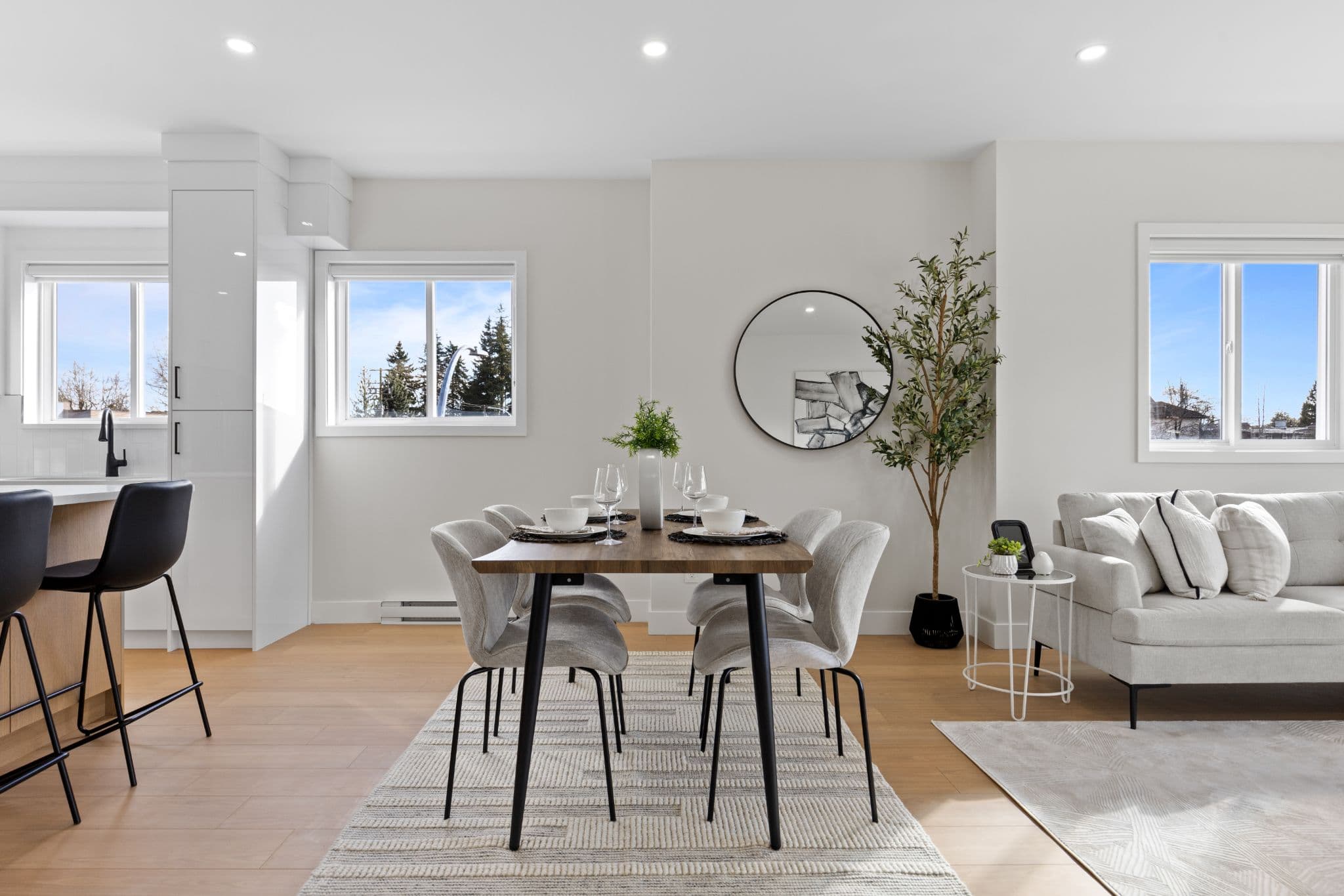 Clearbrook Commons – Dining table set for six with neutral fabric chairs, patterned rug, and circular mirror on the wall next to living room seating.