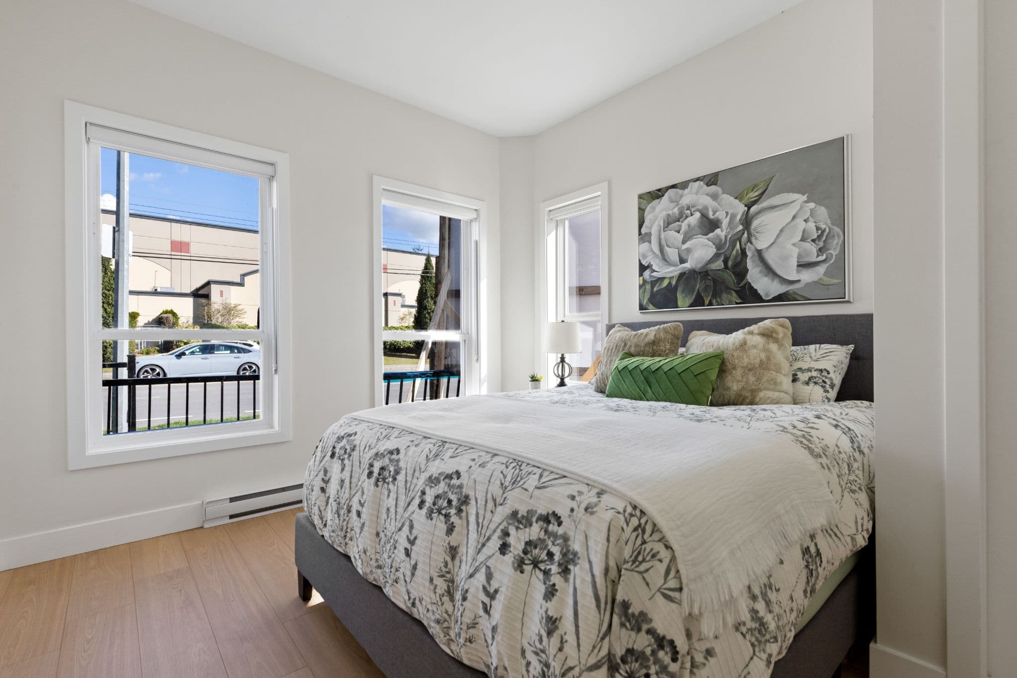Clearbrook Commons – Bedroom with upholstered bed frame, floral print duvet, green accent pillow, and large windows.