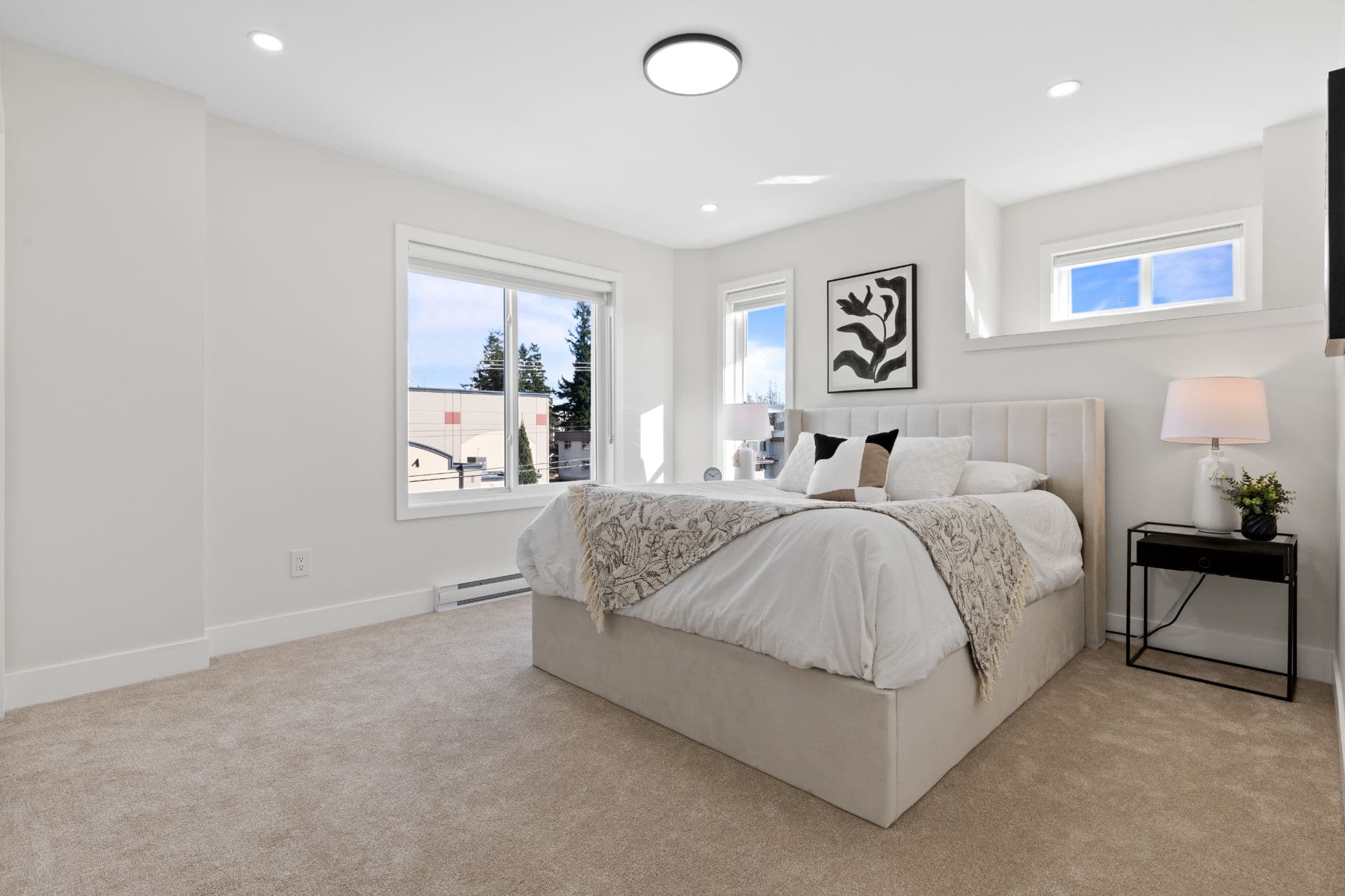 Clearbrook Commons – Spacious bedroom with carpeted flooring, double windows, bed with upholstered headboard, and bedside table with lamp.