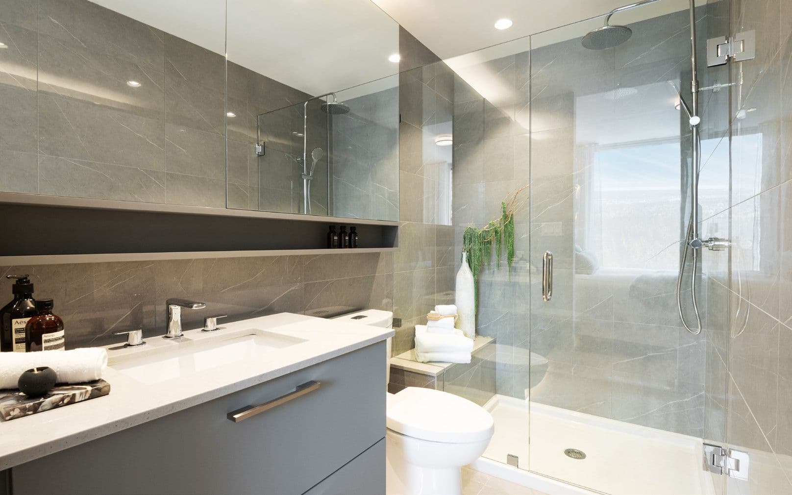 Perla Condos – A bathroom with a grey stone-tiled glass shower and soaker tub, a floating vanity with dark cabinetry, and a window looking out over the city.