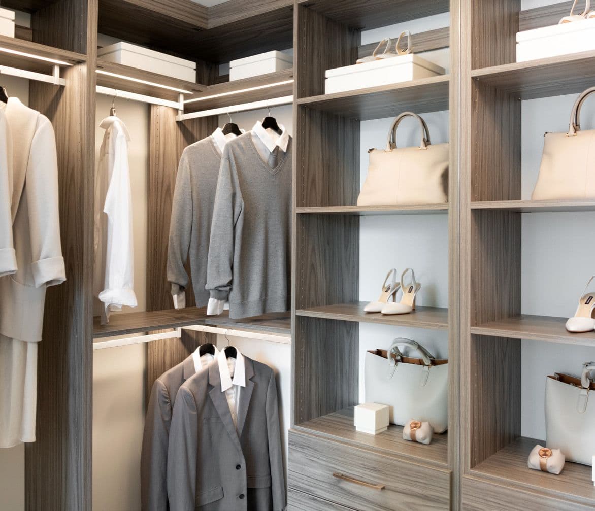 Perla Condos – A walk-in closet with wood shelving displaying folded sweaters, hanging shirts and a blazer, and shelves of handbags and shoes.