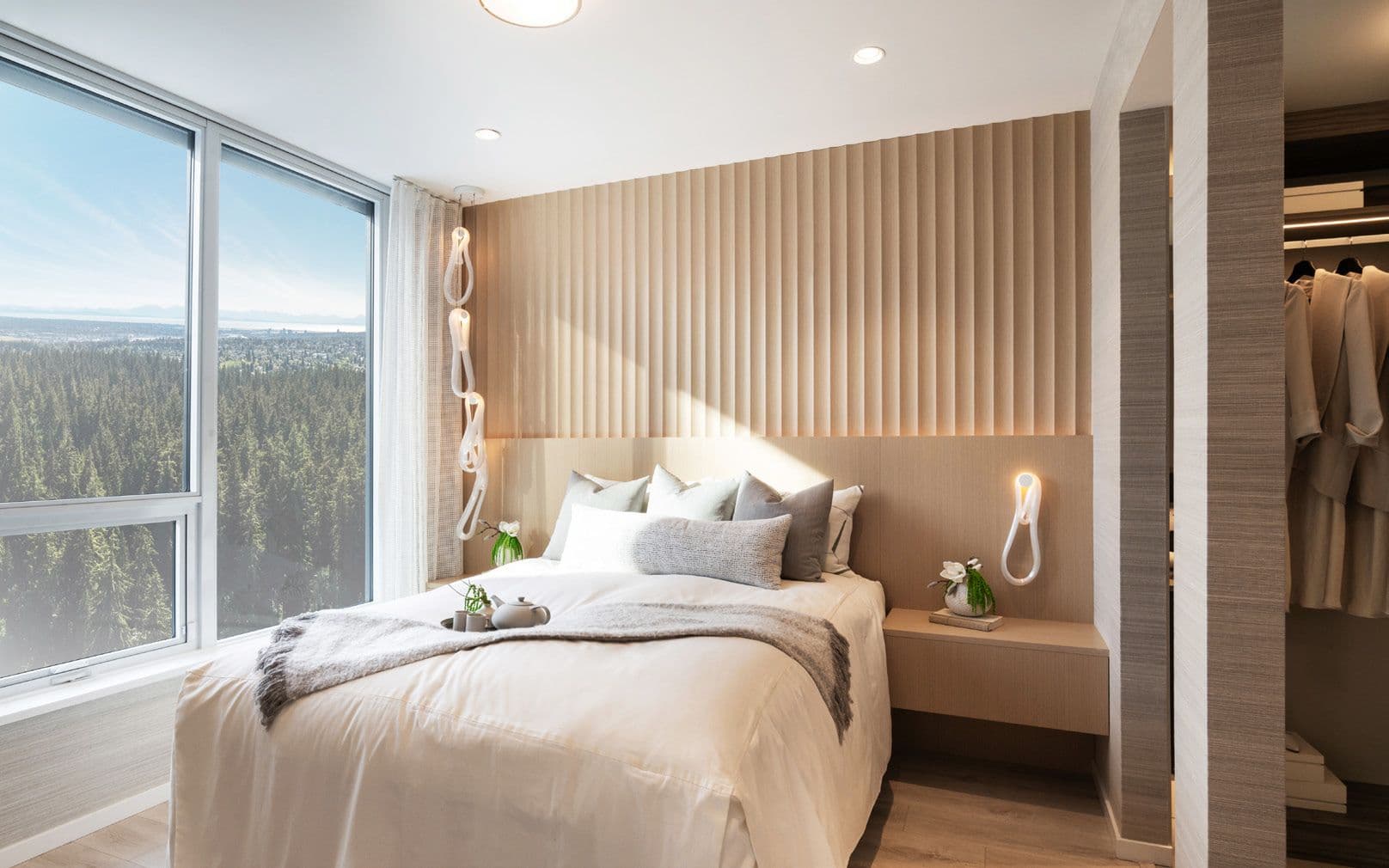 Perla Condos – A bedroom with a white bed and wood slat headboard, a walk-in closet visible to the side, and a floor-to-ceiling window overlooking forest and city in the distance.