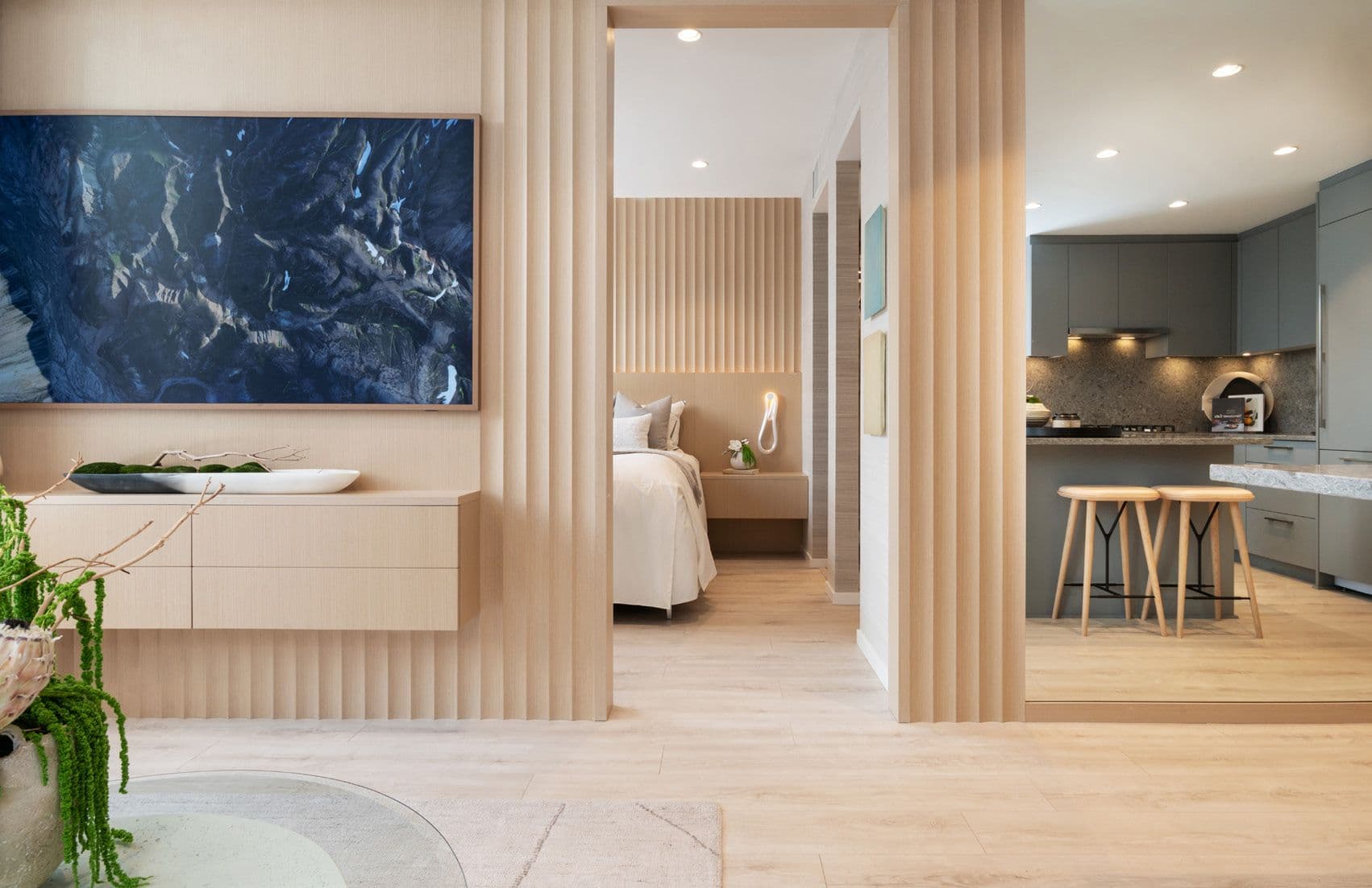 Perla Condos – An open-concept condo interior with a wood-slatted feature wall and wall-mounted TV, opening onto a bedroom on one side and a grey kitchen with bar stools on the other.