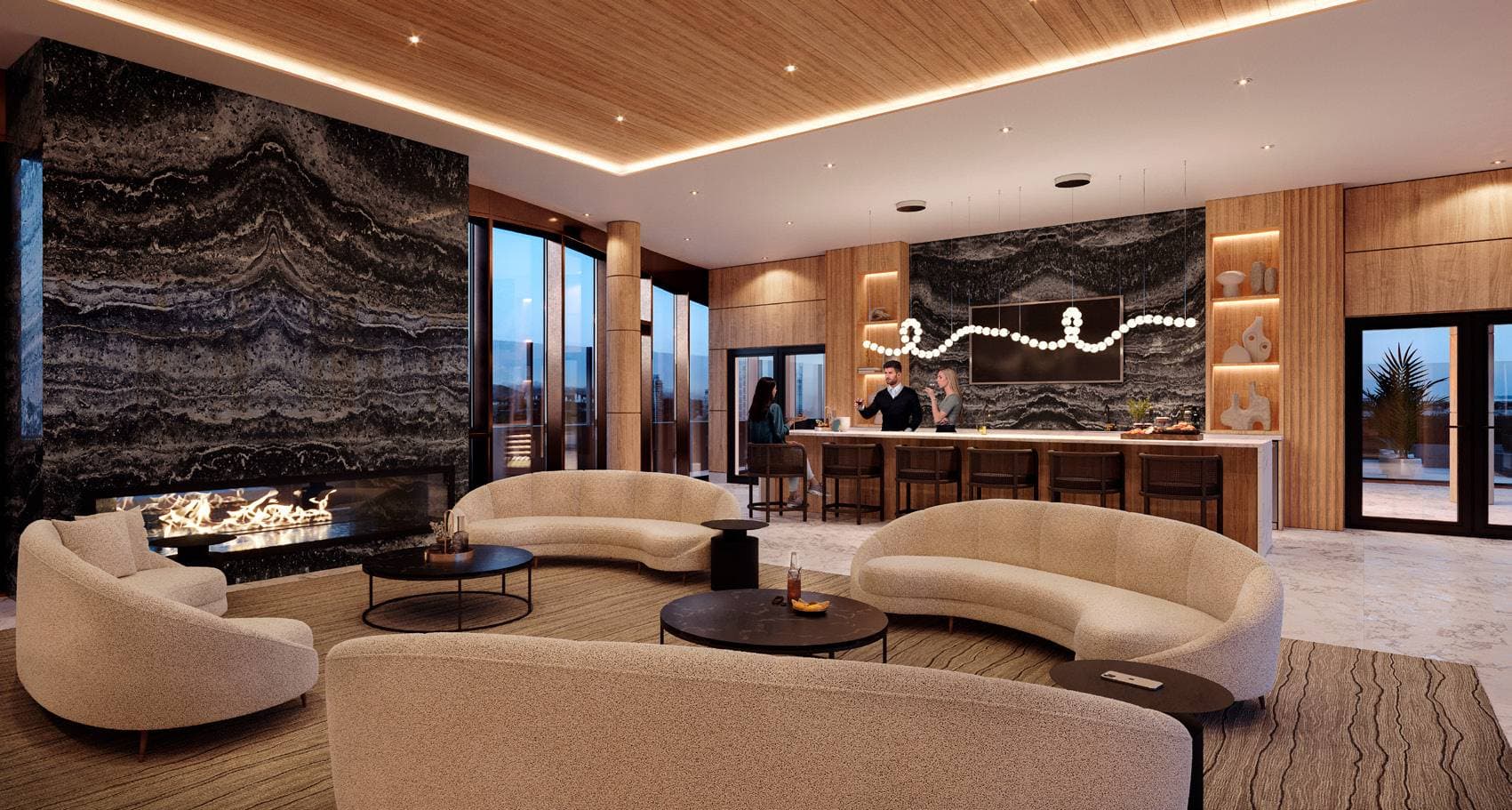 Perla Condos – A resident lounge with a large stone fireplace feature wall, curved beige sofas around coffee tables, and a bar with people seated on stools in the background.