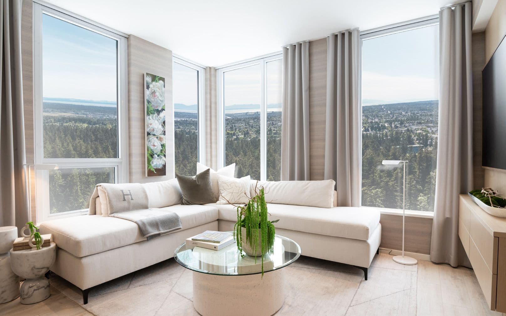 Perla Condos – A living room with a white L-shaped sectional sofa and glass coffee table, wrapped in floor-to-ceiling windows showing a sweeping view of forest, city, and distant mountains.