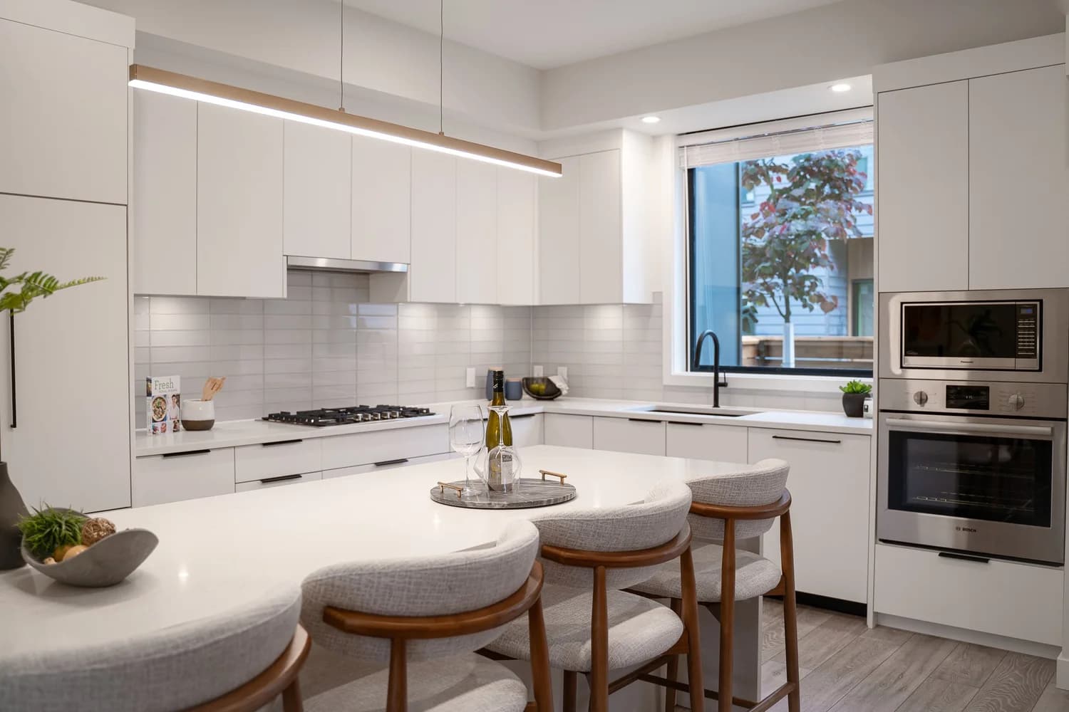 Brighouse 22 – A modern, bright kitchen featuring flat-panel white cabinetry, white quartz countertops, a matching backsplash, and a large island with three cushioned wood-framed stools. Stainless steel appliances, including a built-in wall oven, gas cooktop, and a linear pendant light above the island, complete the clean aesthetic.