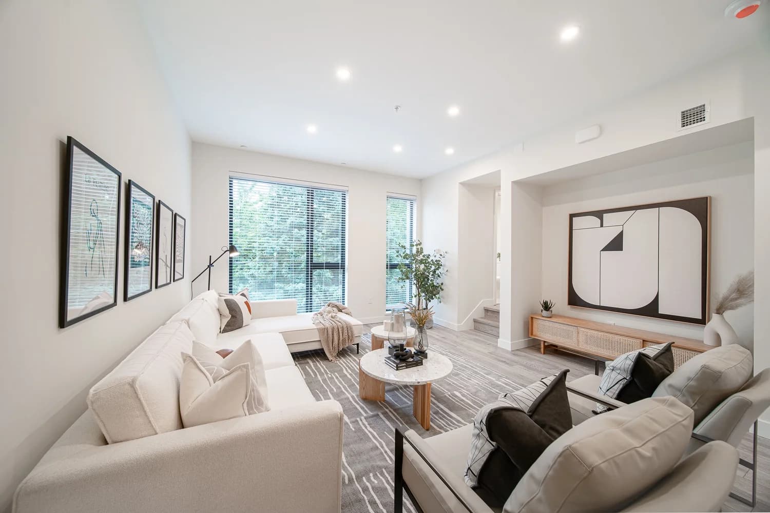 Brighouse 22 – A modern, bright living room features a large cream-colored sectional sofa paired with two light grey armchairs arranged around round marble coffee tables on a patterned area rug. Large black-framed windows with blinds reveal lush outdoor greenery, while framed abstract artwork adorns the neutral white walls.