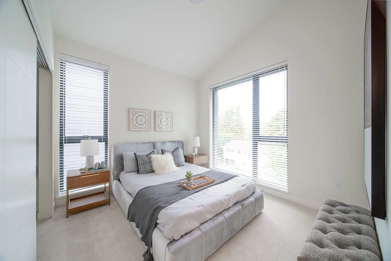 Brighouse 22 – A modern, bright bedroom features a plush grey upholstered bed staged with decorative pillows, a wooden tray, and a dark grey throw blanket. Bright natural light fills the space through two large windows fitted with horizontal blinds, set against off-white walls and neutral carpeting.