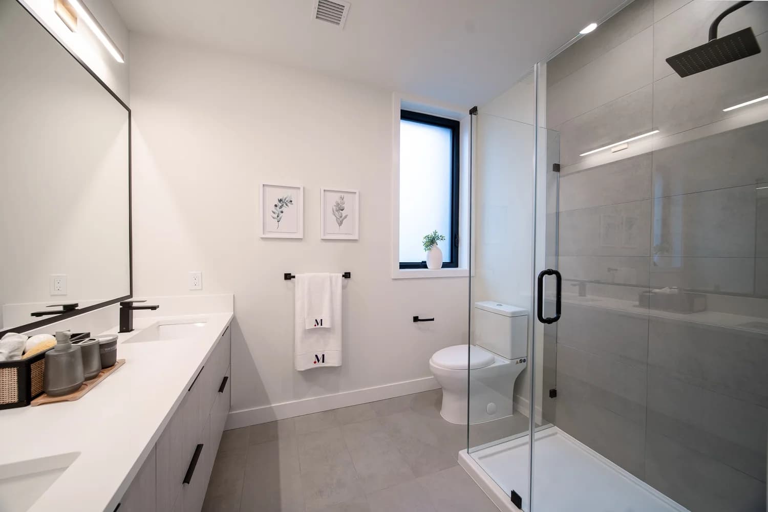 Brighouse 22 – A modern bathroom featuring a double-sink vanity with white quartz countertops, light wood cabinetry, and sleek matte black hardware. A frameless glass walk-in shower with large-format grey tile walls and a square rainfall showerhead sits beside a white toilet and a frosted window.