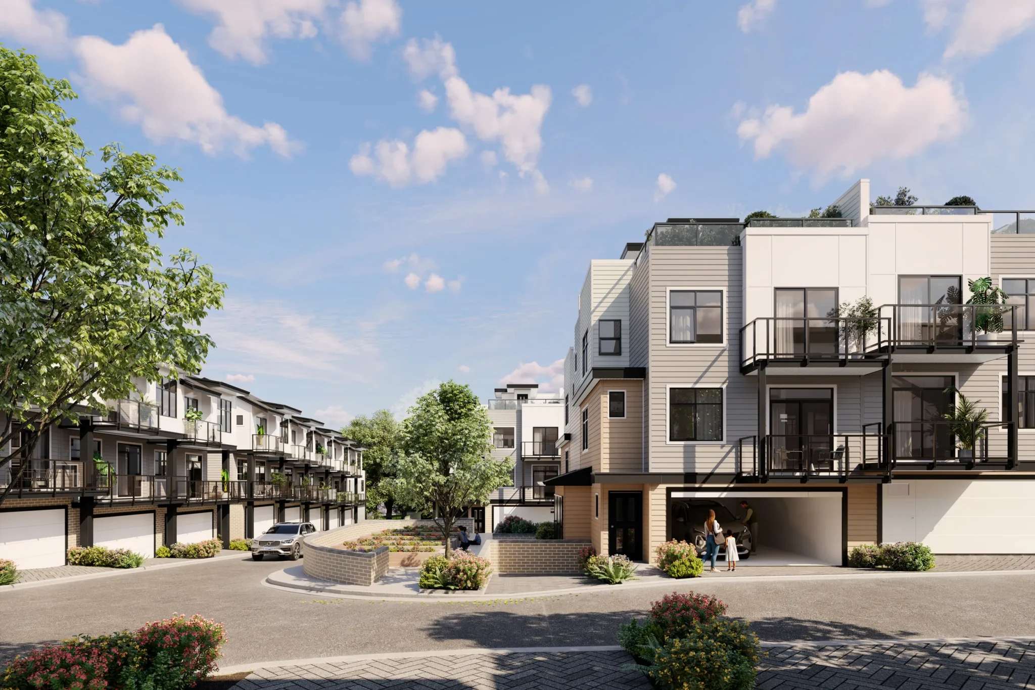 Goldstone – A ground-level view of a contemporary townhouse community featuring multi-story units with light horizontal siding, black-framed windows, and private balconies with glass railings. An internal paved roadway winds past integrated ground-floor garages, small brick retaining walls, and landscaped greenery under a bright, partly cloudy daytime sky.