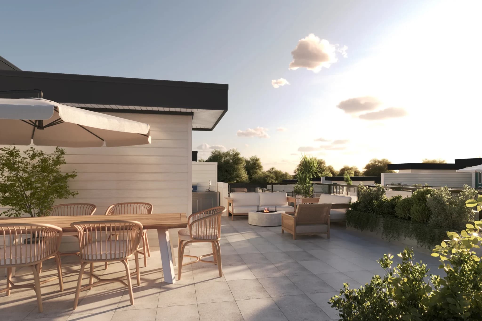 Goldstone – A sunlit rooftop terrace features a tiled patio furnished with a wooden dining table under an umbrella and an outdoor lounge set around a fire pit. Planters with lush greenery border the glass railings, overlooking surrounding treetops during golden hour.