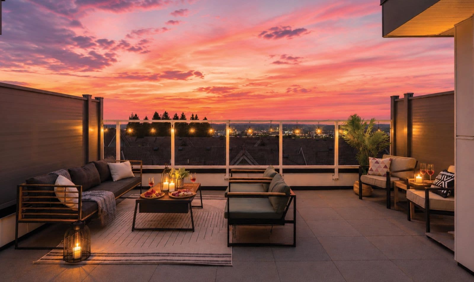 Rising Sun – A spacious rooftop patio terrace furnished with modern outdoor sofas, armchairs, an area rug, and a coffee table arranged with wine and charcuterie. String lights hang along the glass railing, set against a dramatic, vibrant pink and orange sunset over distant rooftops and treetops.