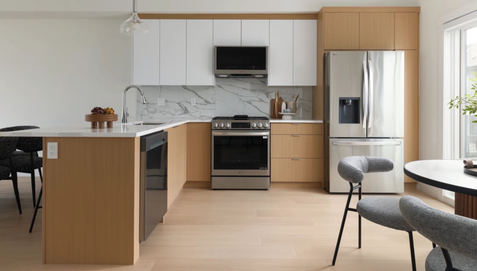 Rising Sun – A modern open-concept kitchen features light natural wood lower cabinetry, flat-panel white upper cabinets, and a white marble-patterned backsplash and countertop. Stainless steel appliances include a slide-in gas range, built-in microwave, French-door refrigerator, and dishwasher, set against pale hardwood flooring with dining chairs visible in the foreground.