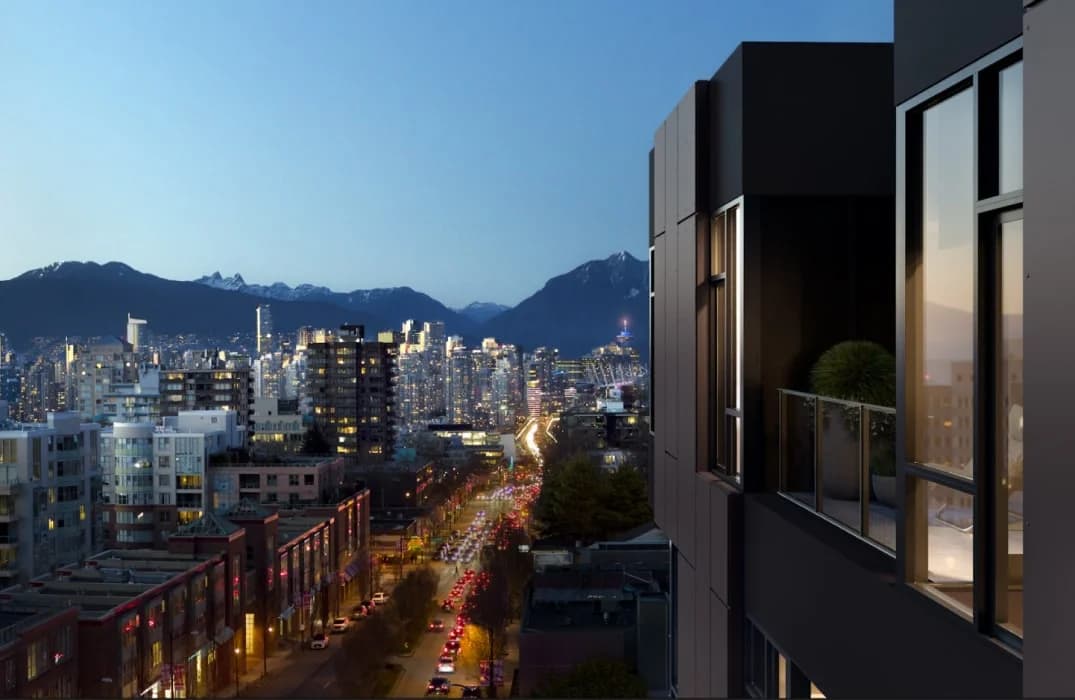 W16 by Olson Kundig – An elevated twilight shot looking past dark, modern matte-black exterior panels and a private balcony toward a vibrant city skyline. Headlights blur along a busy street below, with dense high-rises and dark mountain peaks standing out against a deep blue evening sky.