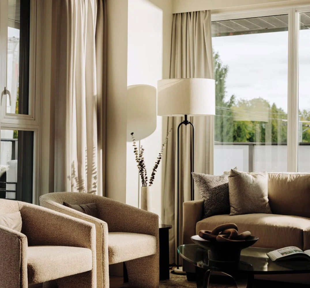 W16 by Olson Kundig – A sunlit residential living room featuring a cozy beige sofa, two textured accent armchairs, and a modern black floor lamp beside floor-to-ceiling windows with neutral drapes. Natural light streams into the space, highlighting warm earth tones, a glass coffee table, and views of lush green trees outside.