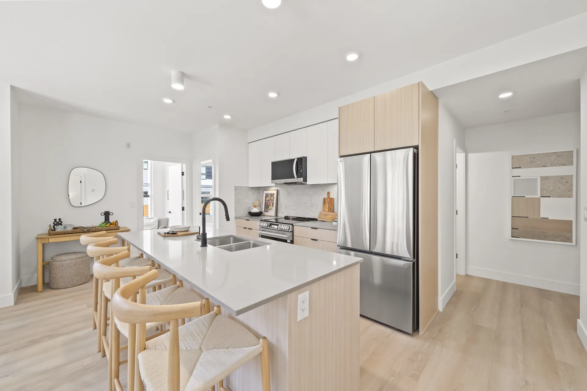 Gabriel – A modern open-concept kitchen features light wood and white flat-panel cabinetry paired with stainless steel appliances and light quartz countertops. A large kitchen island with an undermount sink and matte black faucet is lined with woven-seat wood barstools over light hardwood flooring.