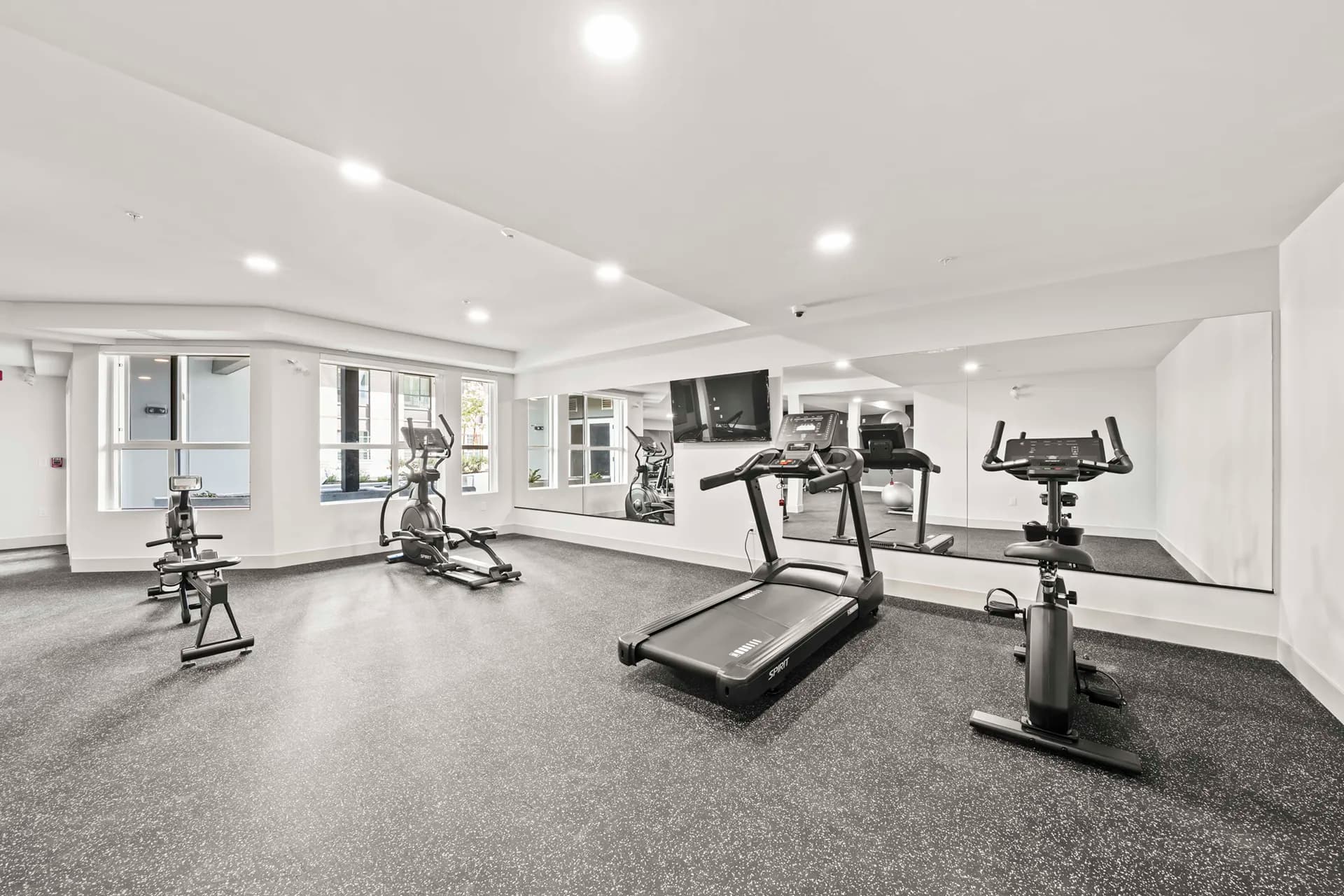 Gabriel – A bright residential fitness room featuring black rubber gym flooring, white walls, and large wall-mounted mirrors reflecting the space. Various cardio equipment, including a treadmill, stationary bike, elliptical, and rowing machine, are arranged in front of windows overlooking an outdoor courtyard.