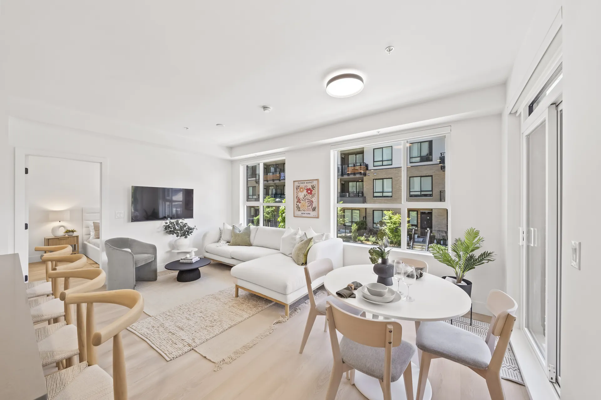 Gabriel – A bright, open-concept living and dining area features light blonde wood flooring, white walls, and large windows looking out onto a residential courtyard. The space is styled with a white sectional sofa, a small circular dining set, woven-seat barstools, and an open doorway leading into an adjacent bedroom.