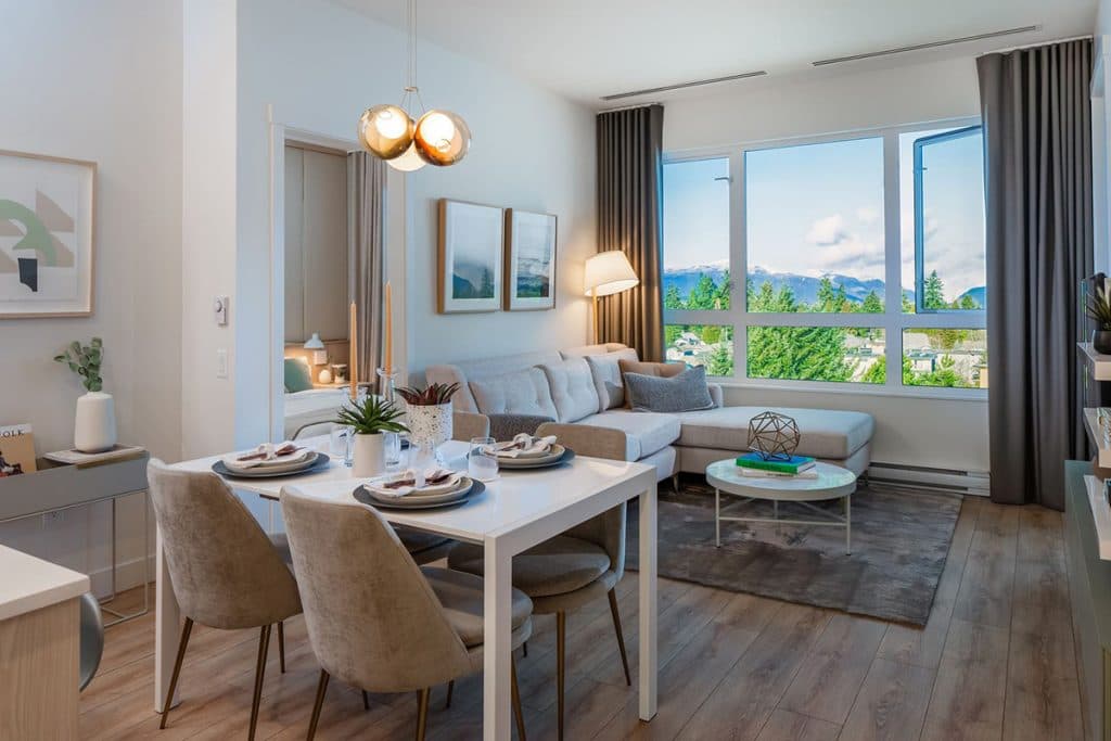 The Oaks Phase 3 (Willow) – A bright living and dining area with a sectional sofa, dining table for four, and large windows looking out to mountains and trees.