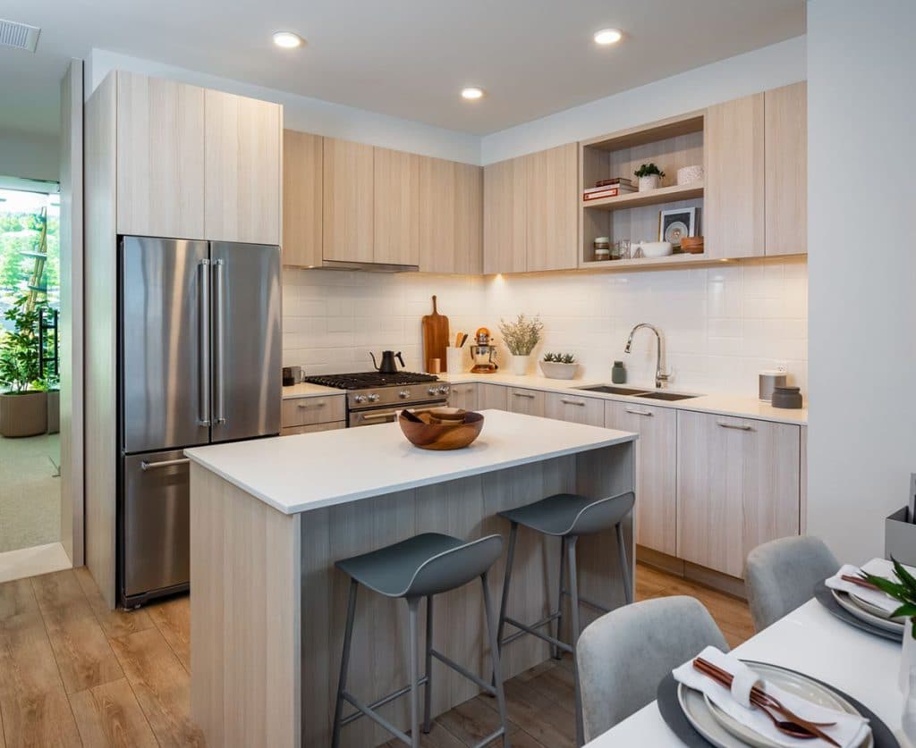 The Oaks Phase 3 (Willow) – A modern kitchen with light wood cabinetry, stainless steel refrigerator and range, and a white waterfall-edge island with bar stools.