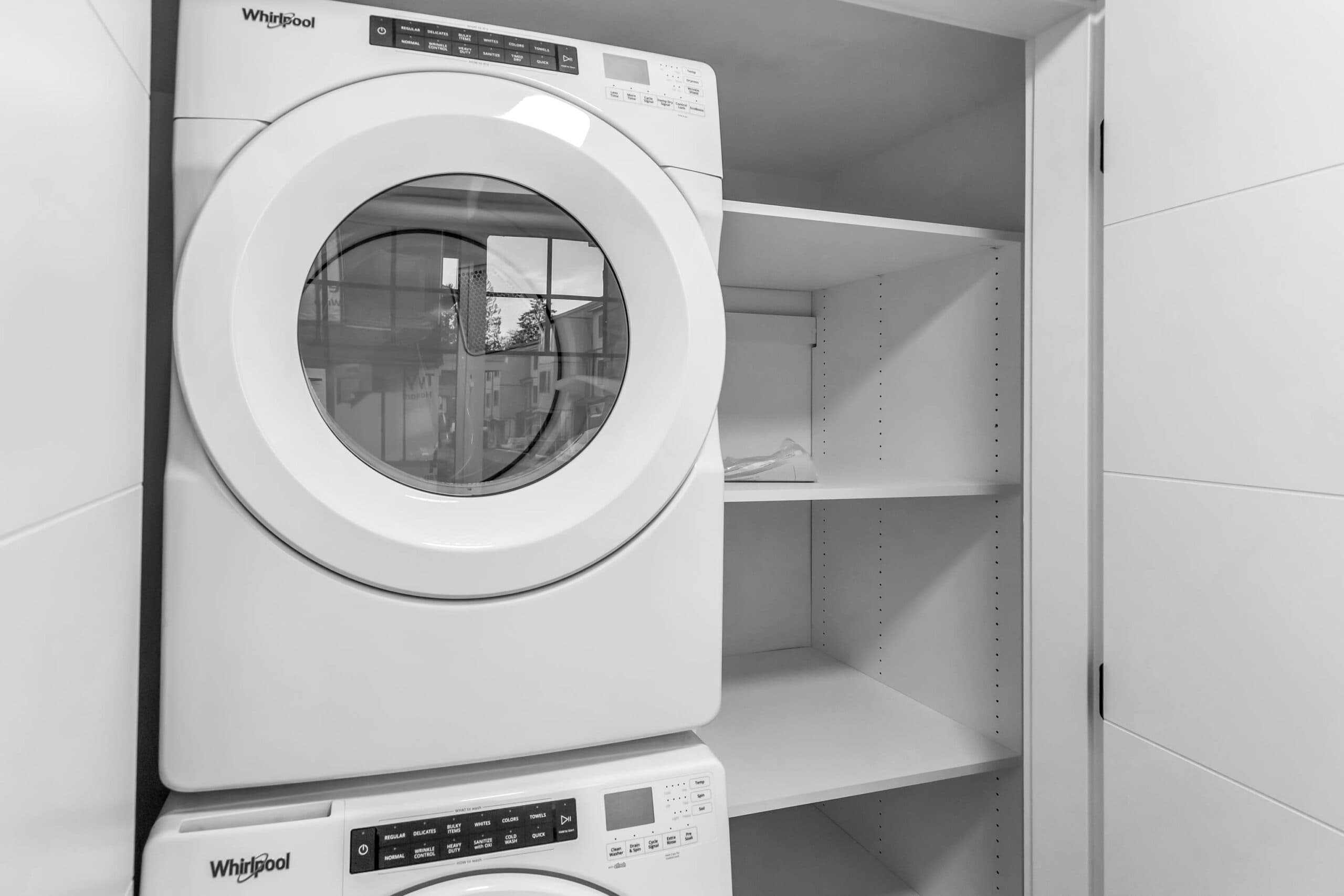 Pine Creek Estates – A white stacked front-loading Whirlpool washer and dryer set fitted neatly inside a bright laundry closet. Adjacent to the machines are built-in adjustable white shelves, creating a functional, compact utility space.