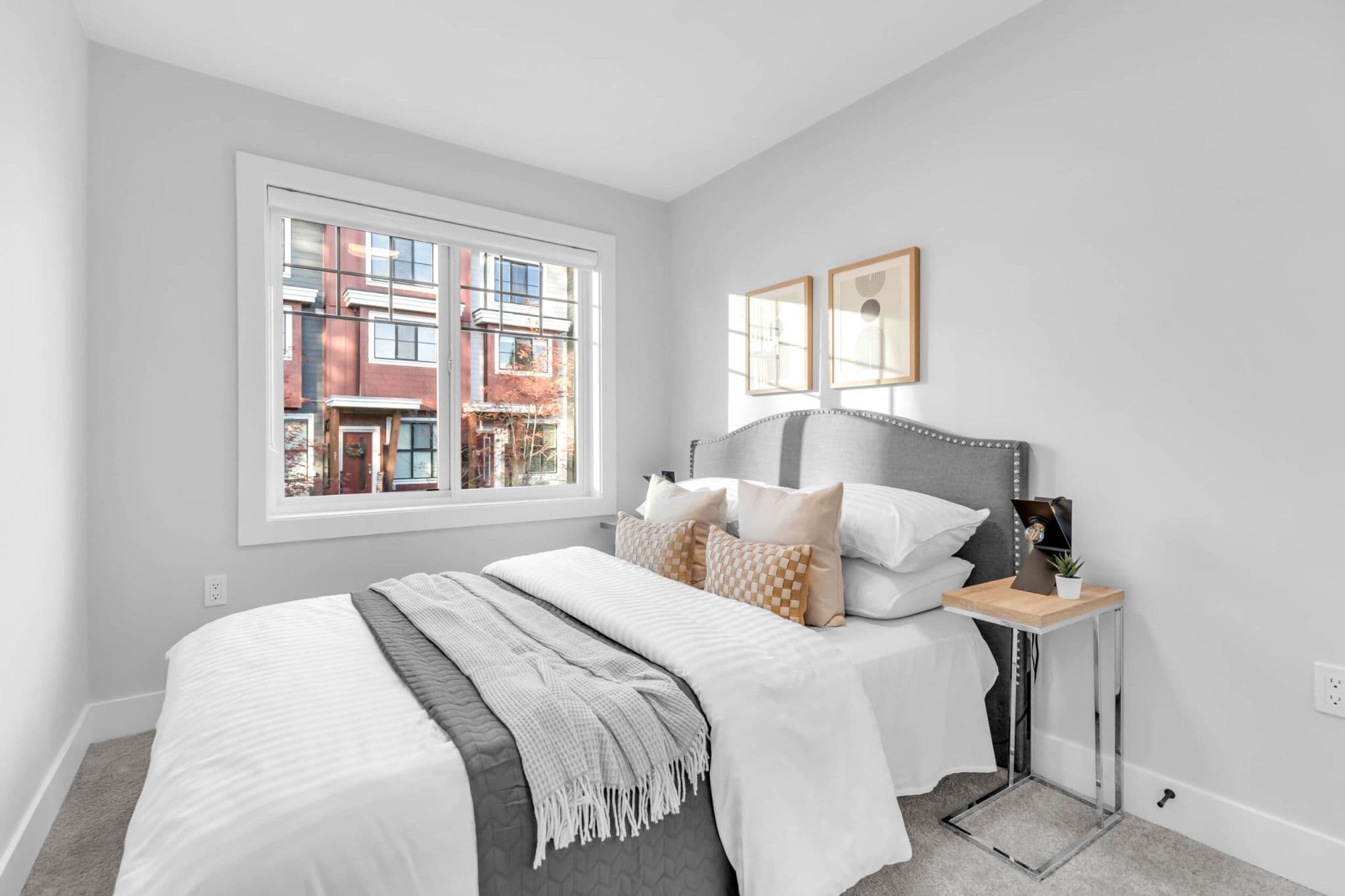 Pine Creek Estates – A bright, modern bedroom featuring a neatly dressed bed with a gray upholstered, nailhead-trimmed headboard and neutral throw pillows. A large window lets in daylight and offers a view of neighboring residential townhomes.