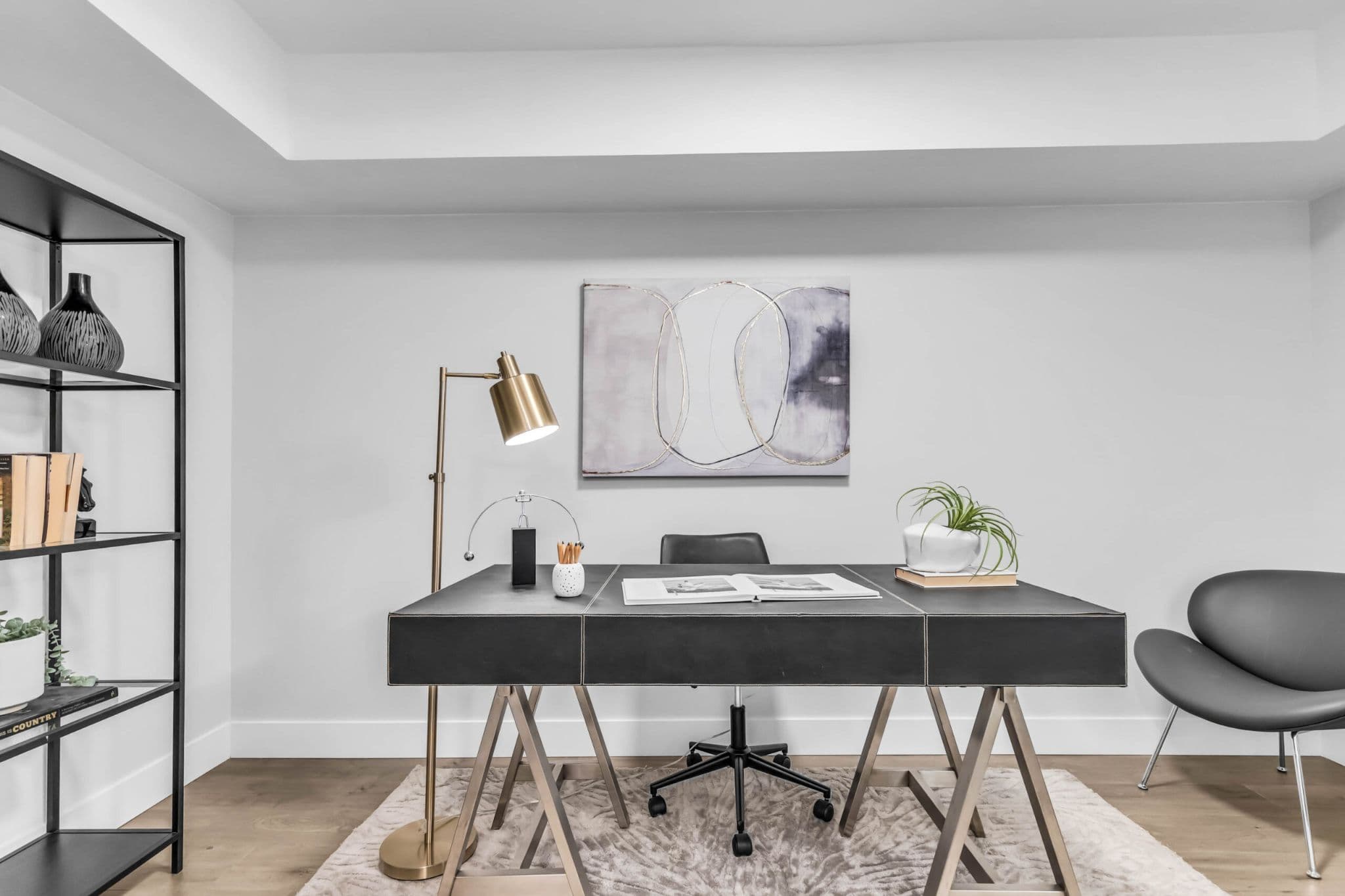 Pine Creek Estates – A modern home office setup featuring a dark leather-style desk with metallic trestle legs centered on a plush neutral area rug. Bright, clean interior lighting illuminates a brass floor lamp, abstract canvas wall art, a open bookshelf, and a charcoal accent chair against crisp white walls.