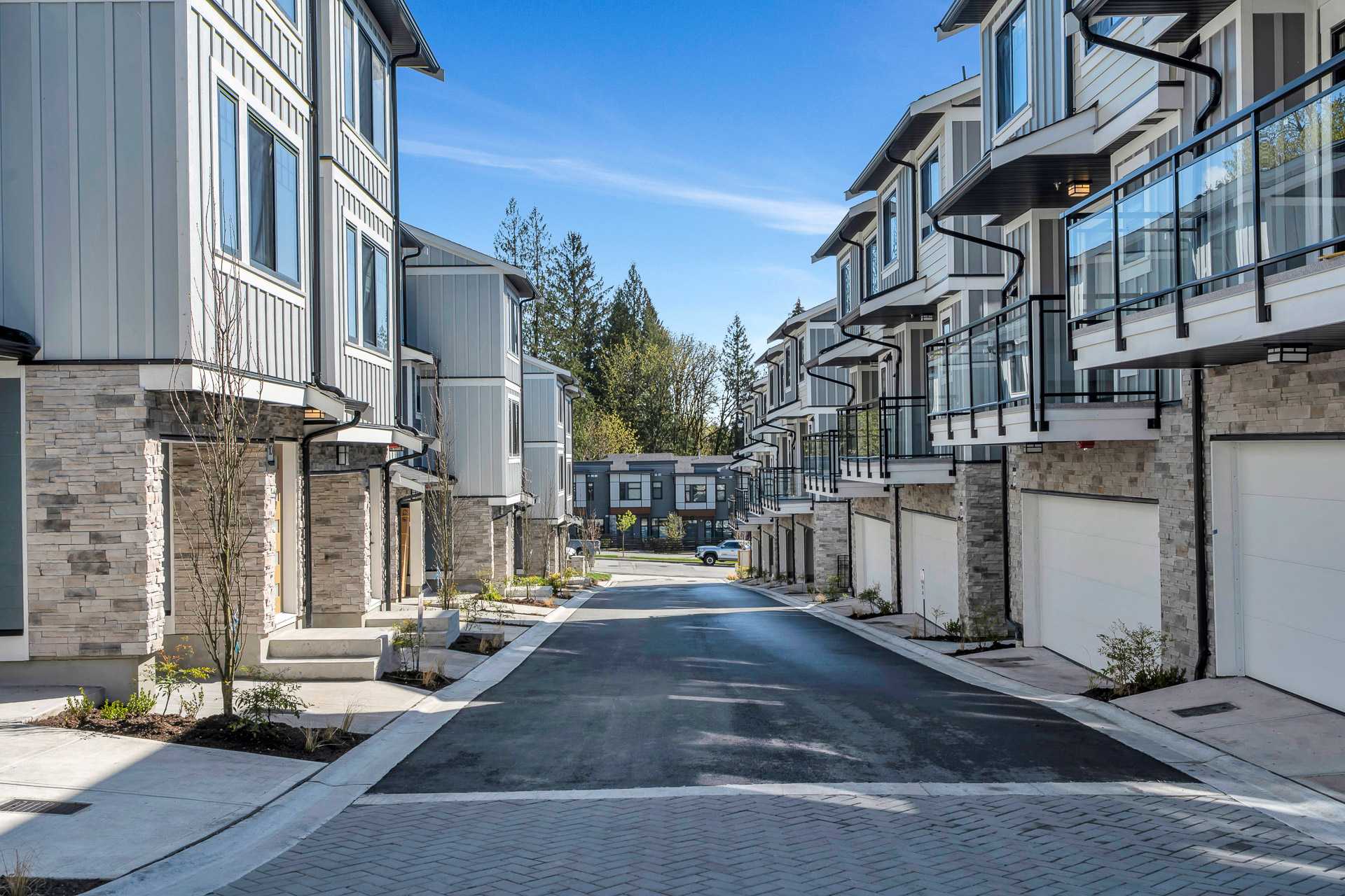 Pine Creek Estates – A ground-level street view looking down a paved internal driveway lined with modern multi-story townhomes. The buildings feature stone veneer bases, light grey board-and-batten siding, black accent trim, white garage doors, and glass-railed upper balconies under a clear blue sky.