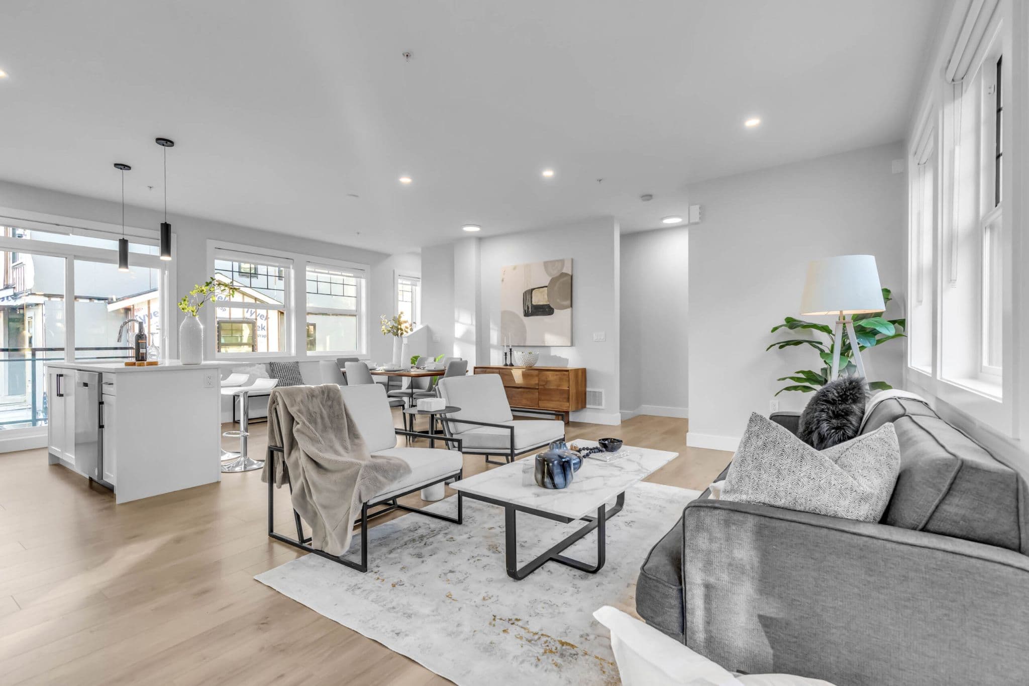 Pine Creek Estates – A bright, open-concept living and dining space featuring light-wood flooring, white walls, and large windows that fill the room with natural light. The area is modernly furnished with a grey fabric sofa, modern armchairs around a marble coffee table, and an adjacent kitchen island with pendant lighting.