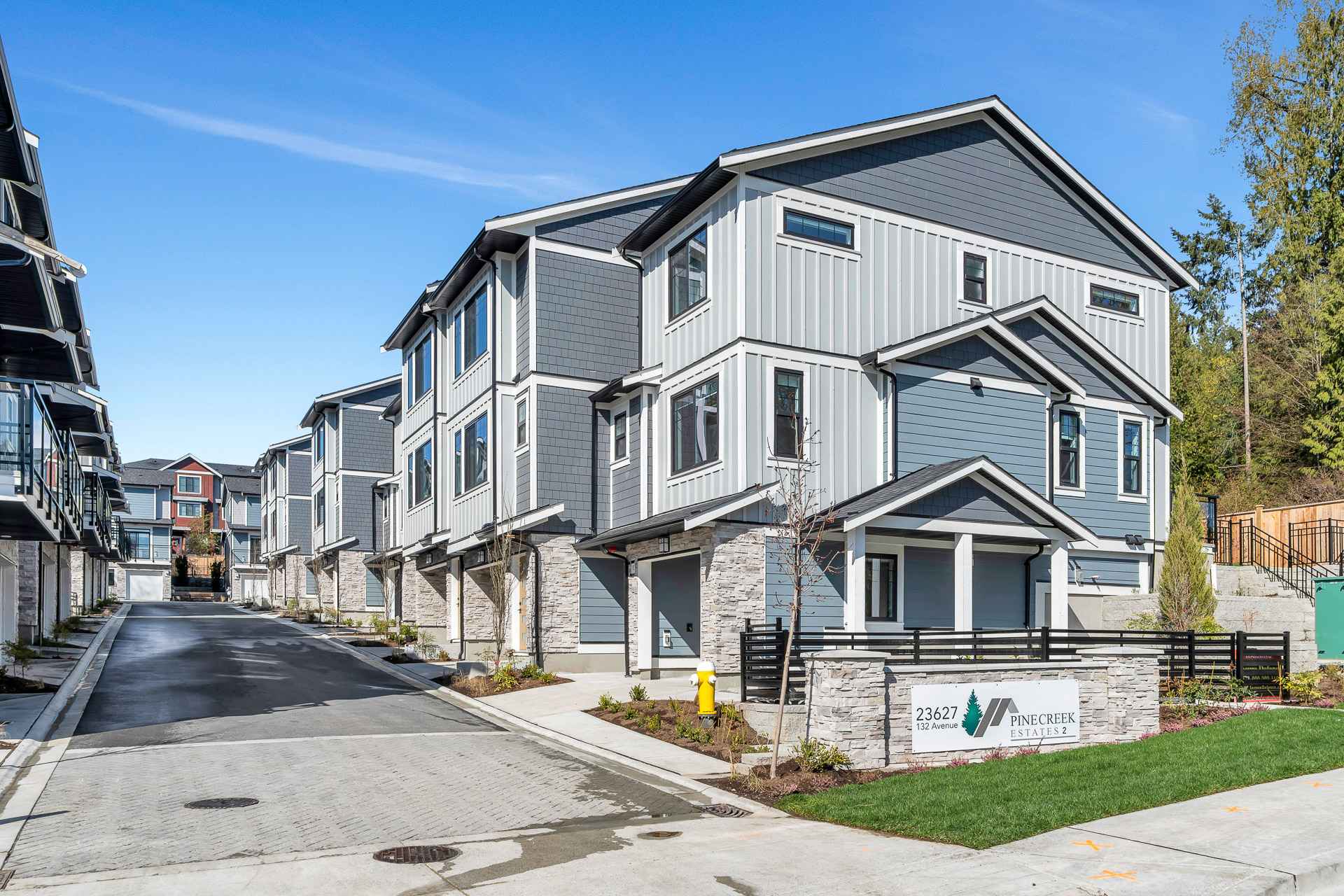 Pine Creek Estates – A ground-level street view of a modern townhome complex featuring blue and light grey siding paired with stone veneer accents along the lower level. An internal paved driveway runs alongside the multi-story buildings under a clear blue sky.