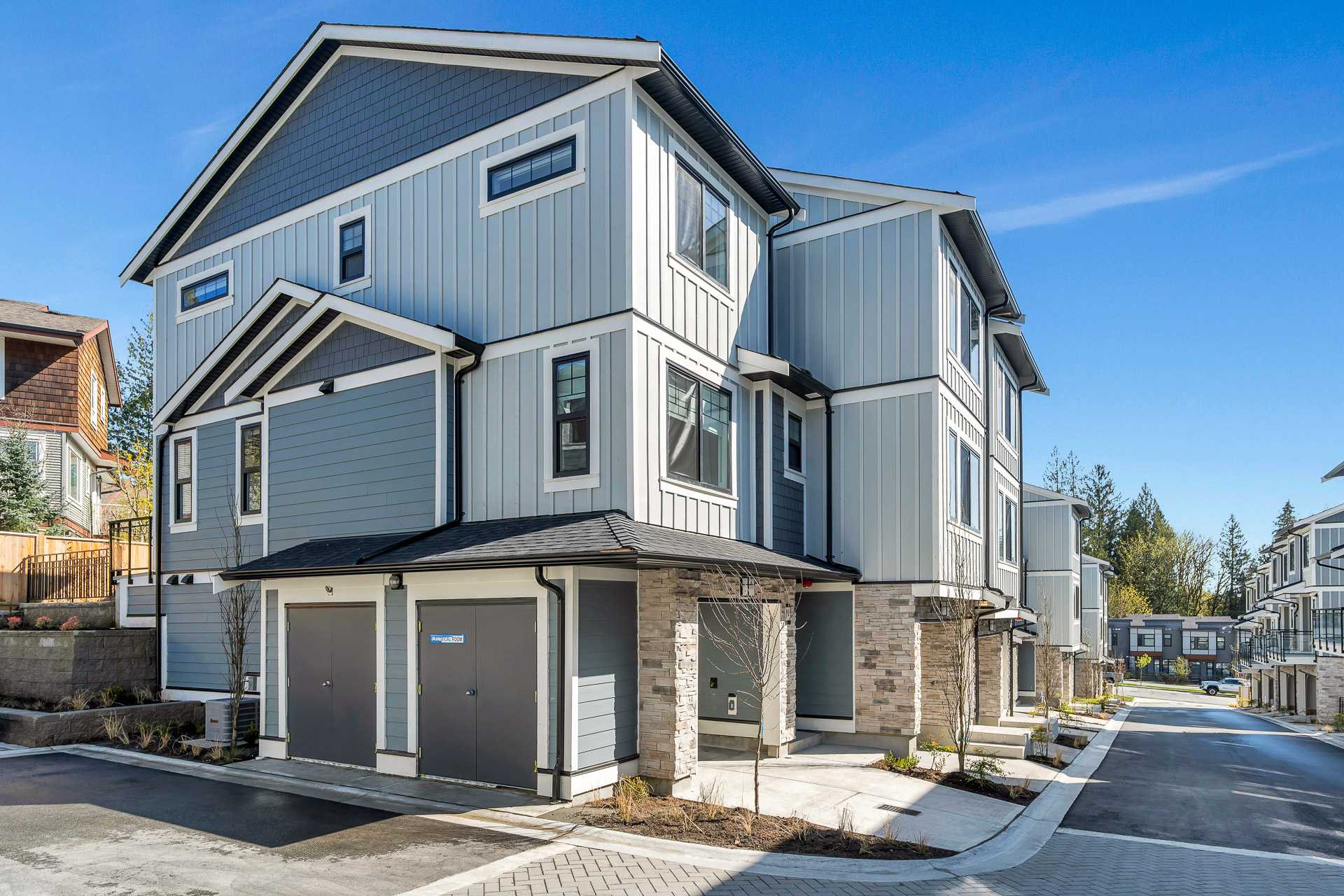 Pine Creek Estates – A ground-level view of a modern multi-story townhouse featuring light grey board-and-batten siding paired with slate-blue horizontal panels and stone accent pillars. The building includes double grey utility doors at the base and sits along a freshly paved residential roadway under a clear blue sky.