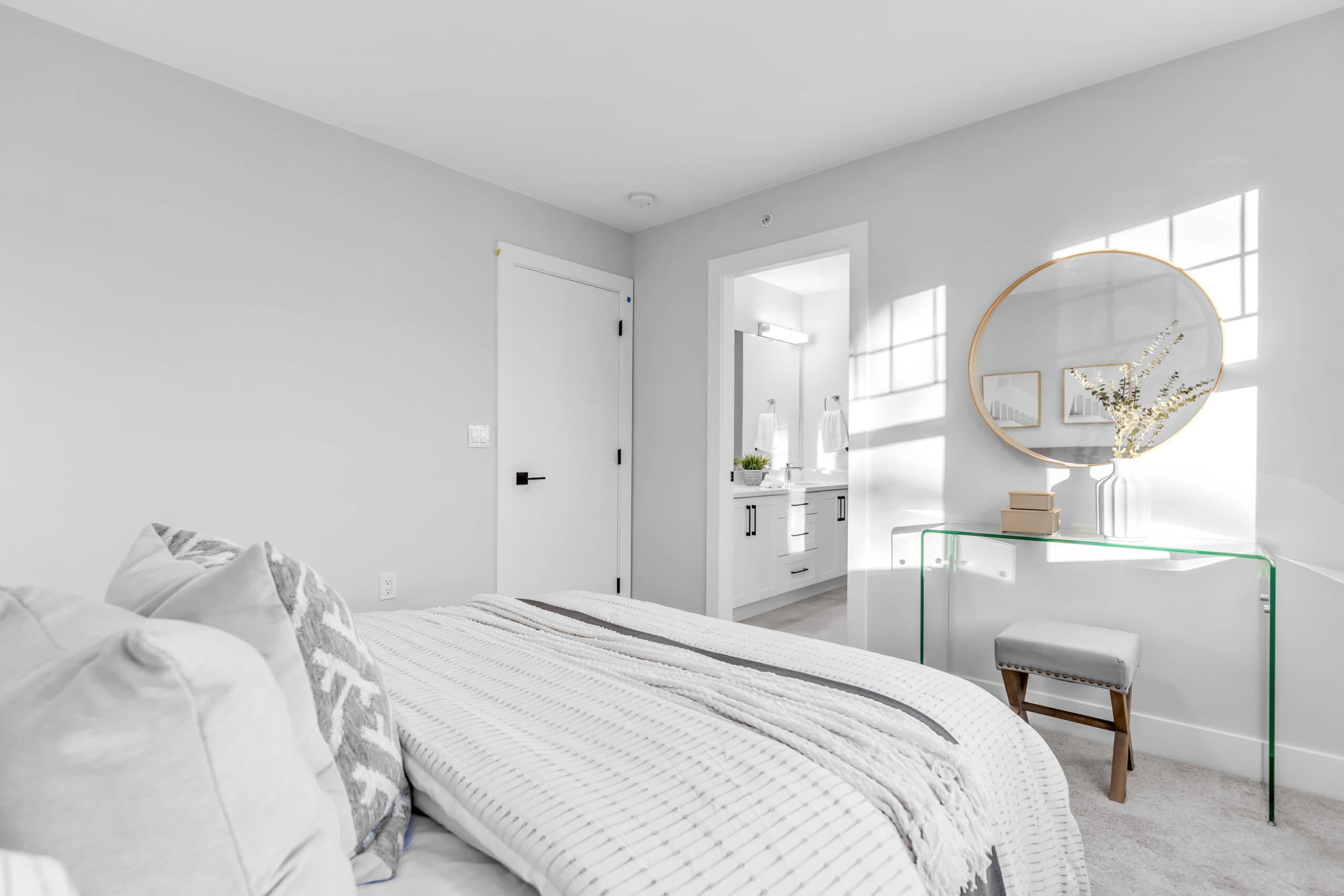 Pine Creek Estates – A bright bedroom with neutral-toned bedding leads into an ensuite bathroom featuring a white vanity. Natural light streams through a window, casting windowpane shadows on the wall above a glass vanity table with a round gold-framed mirror.